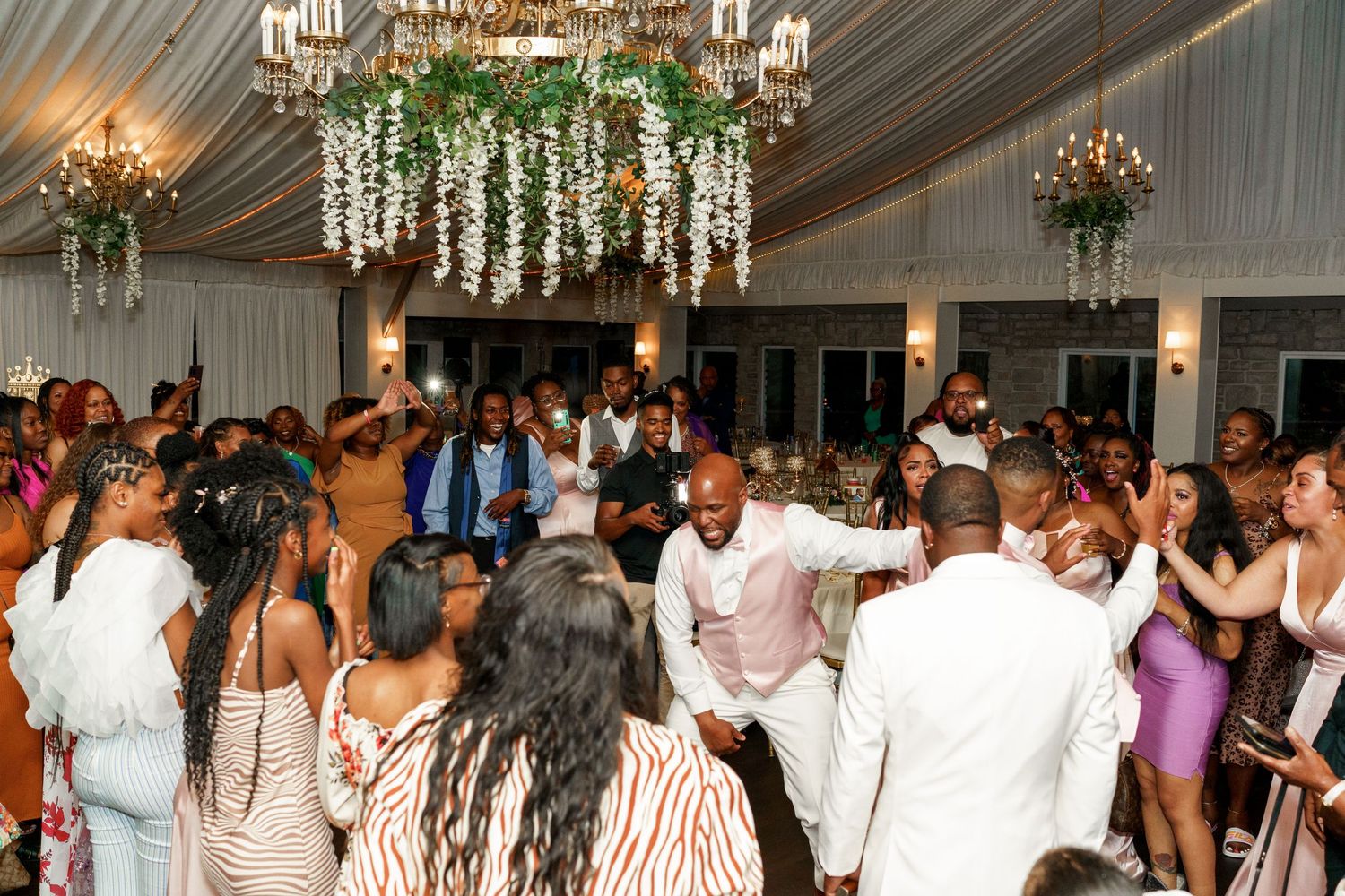 Lively wedding reception dance party under elegant chandeliers and floral decorations.