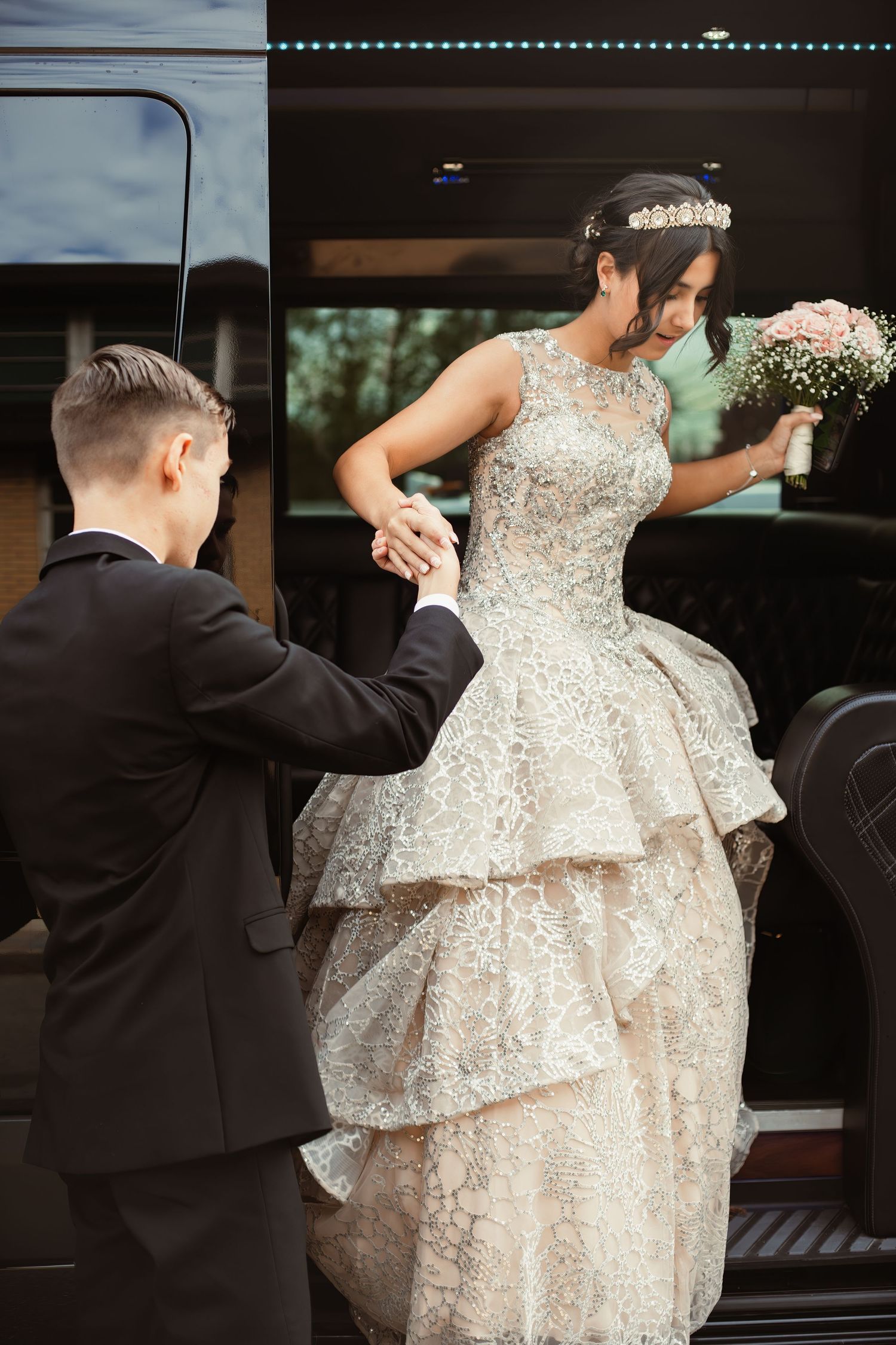 Elegant bride in sparkly gown being assisted from a vehicle while holding flowers.