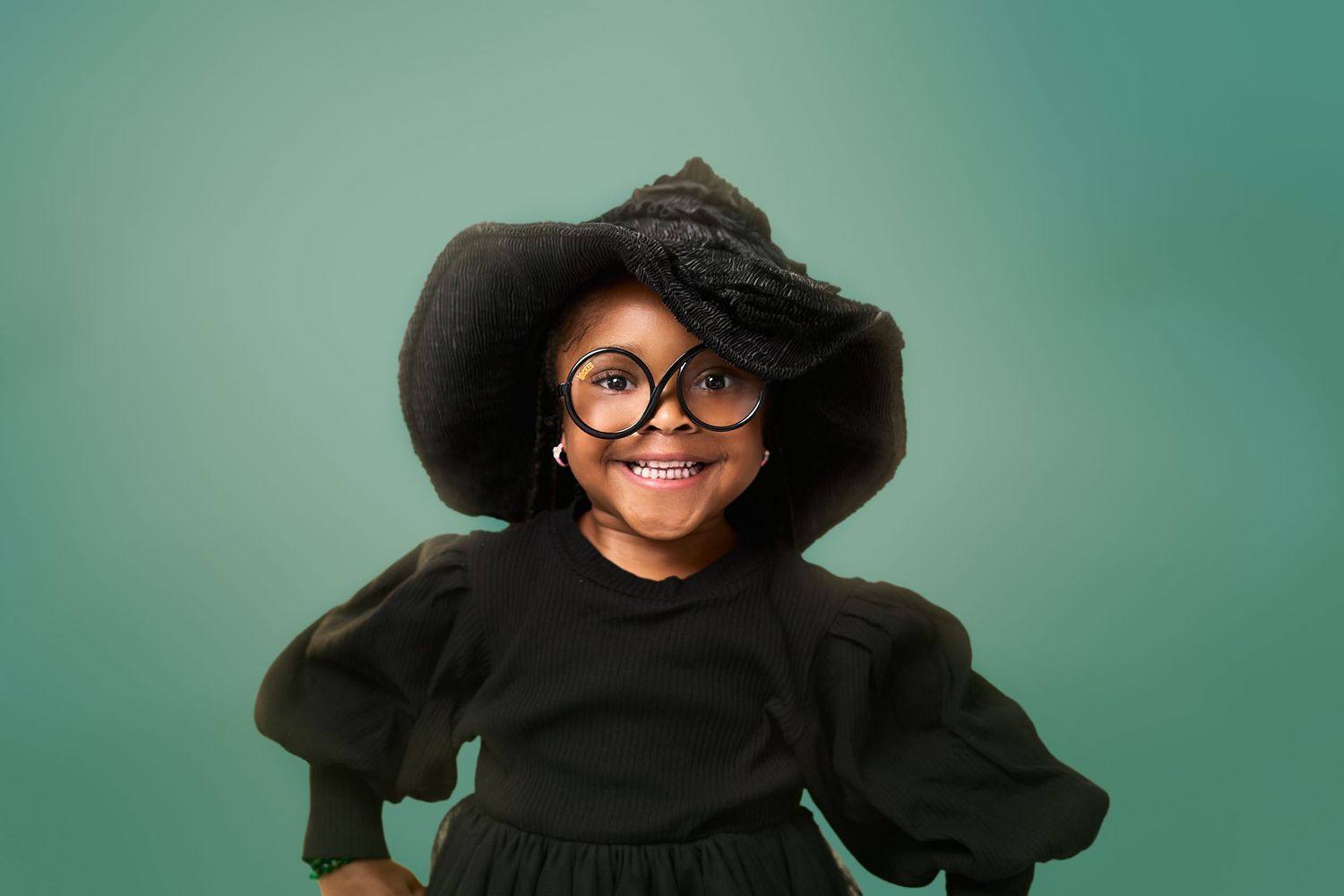 A child in a black dress and wide-brimmed hat wearing round glasses smiles against a green background.