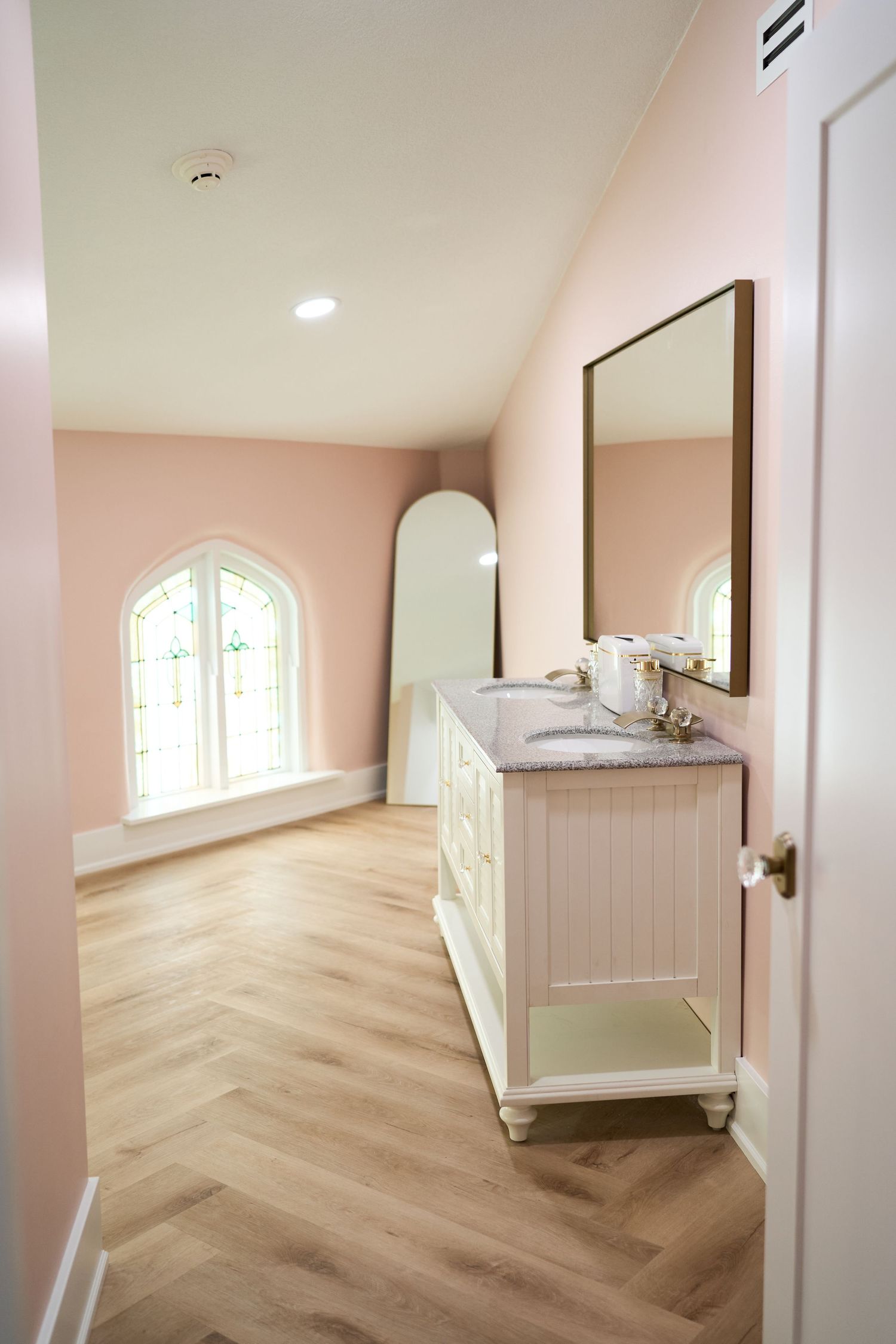 A pink-walled hallway with herringbone wood flooring features an elegant vanity and arched window.