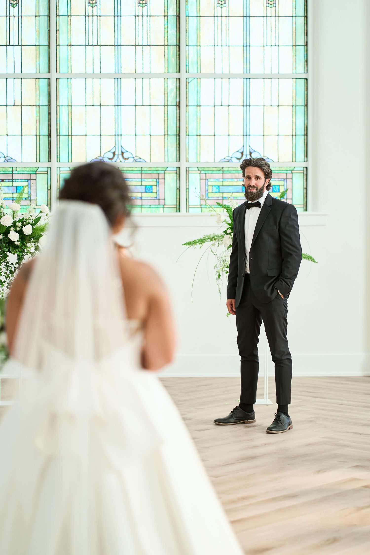 A bride in a white ballgown and groom in black suit share their first look in front of stunning stained glass windows.