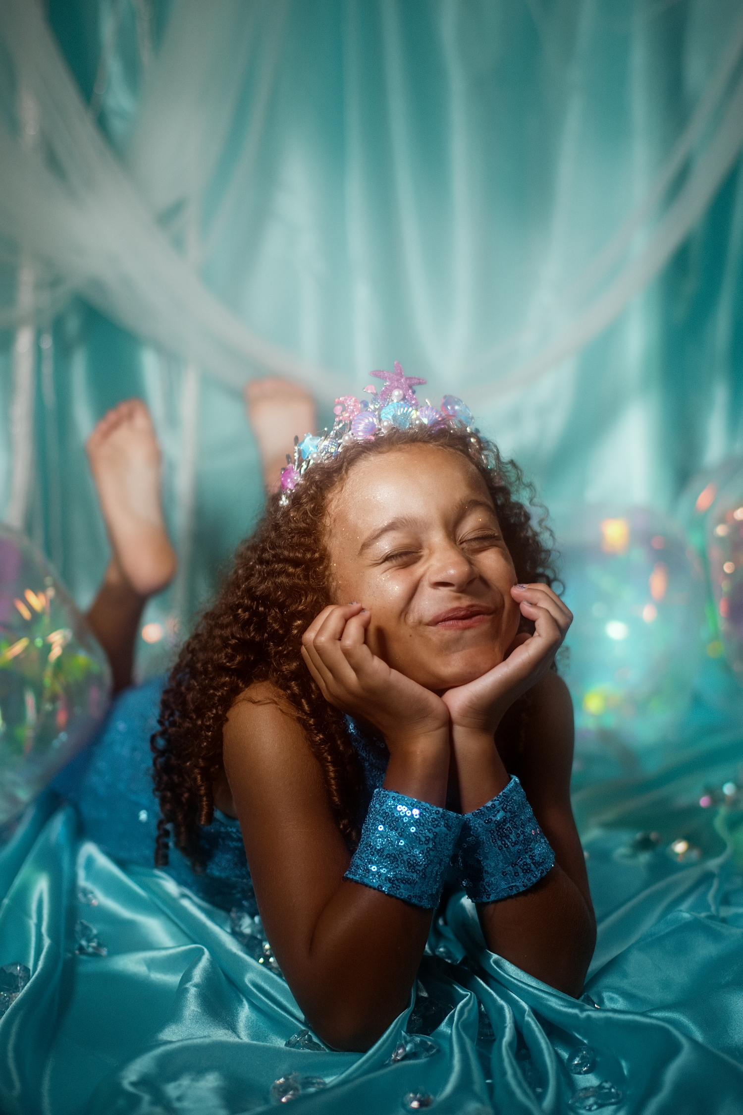 A young girl in a blue sequin dress and crown poses dreamily with hands on cheeks, lying on teal satin fabric.