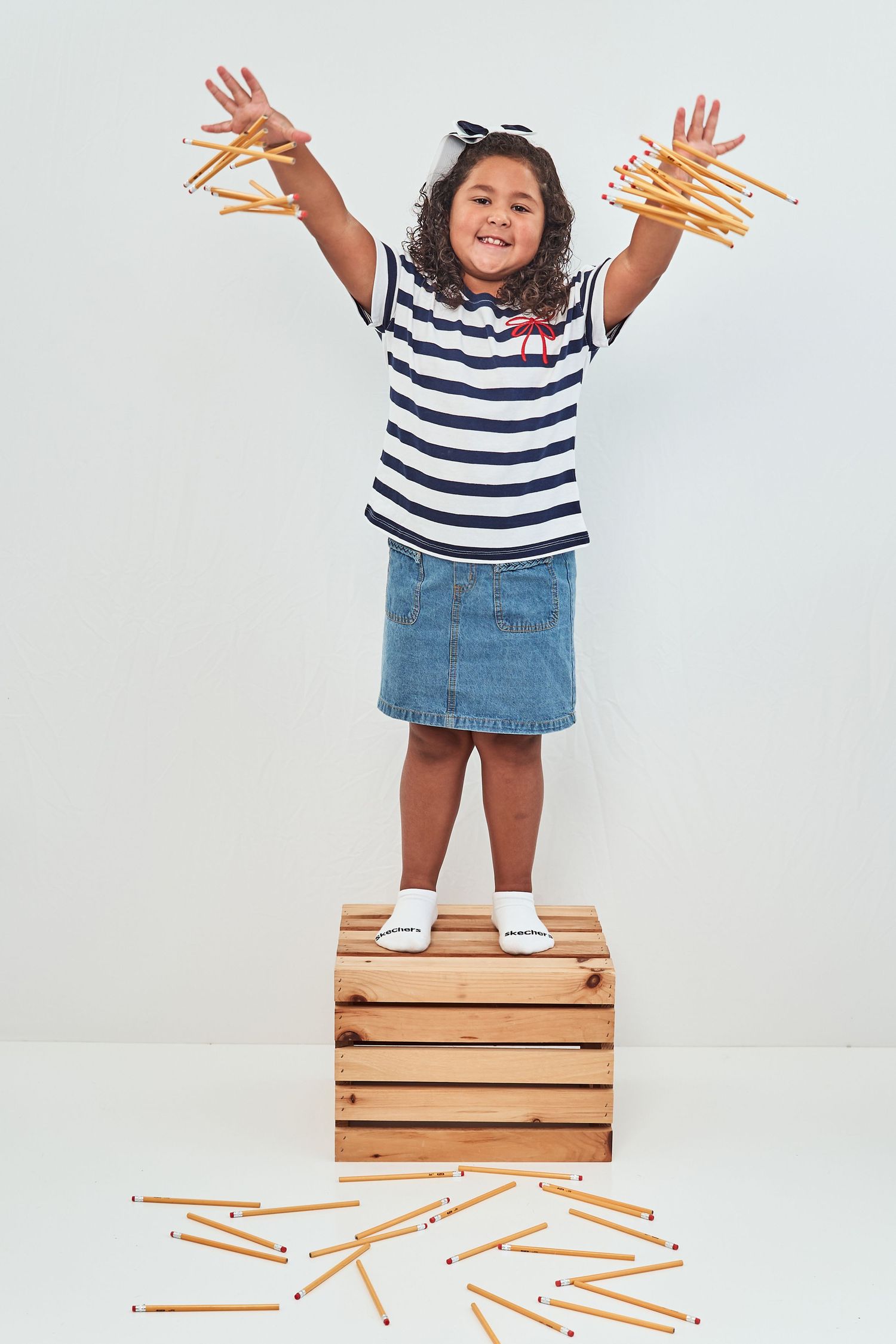 A child in a striped shirt and denim skirt stands on a wooden crate while playfully holding up french fries.