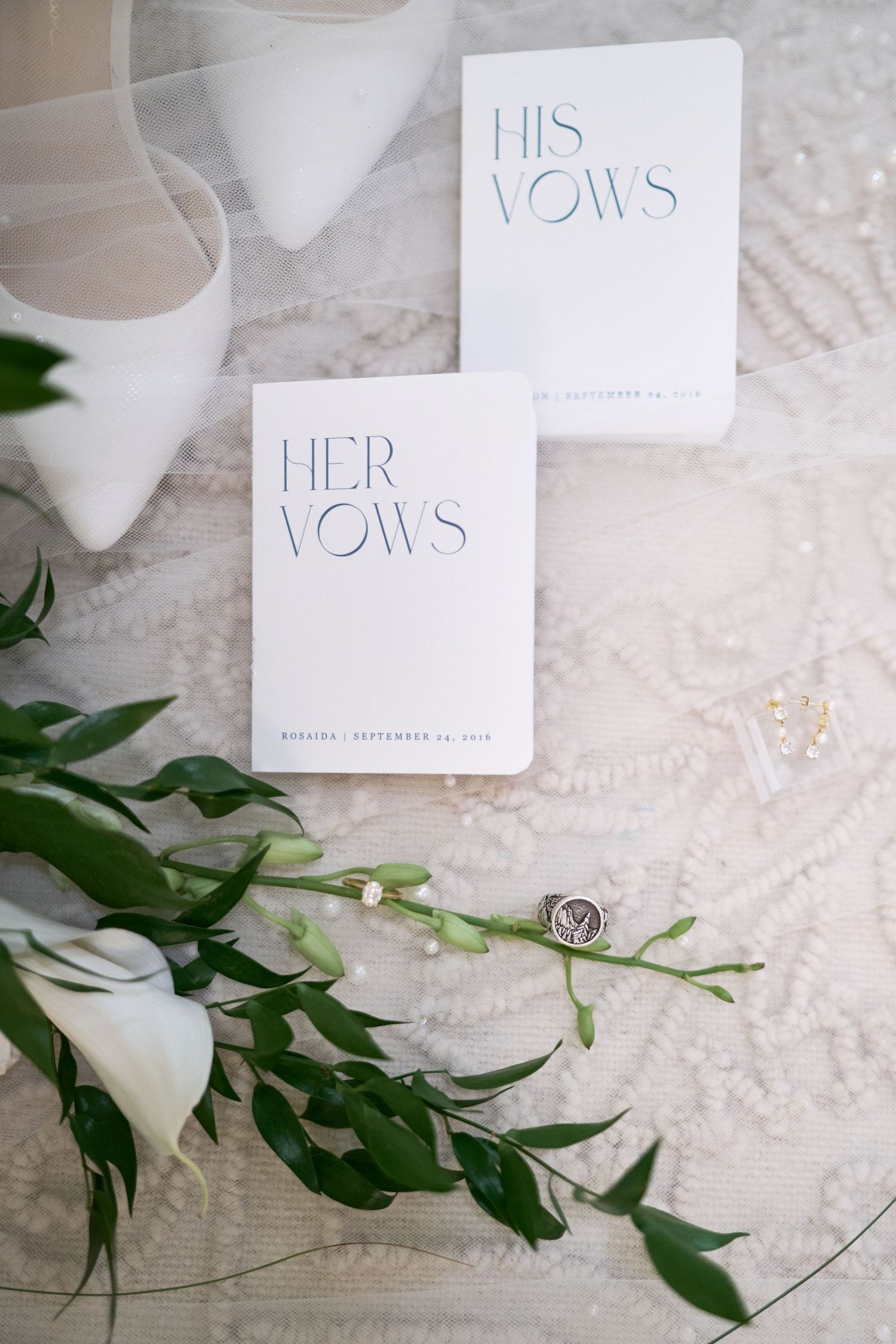 Wedding vow booklets labeled 'His' and 'Her' vows displayed with green foliage on white lace background.
