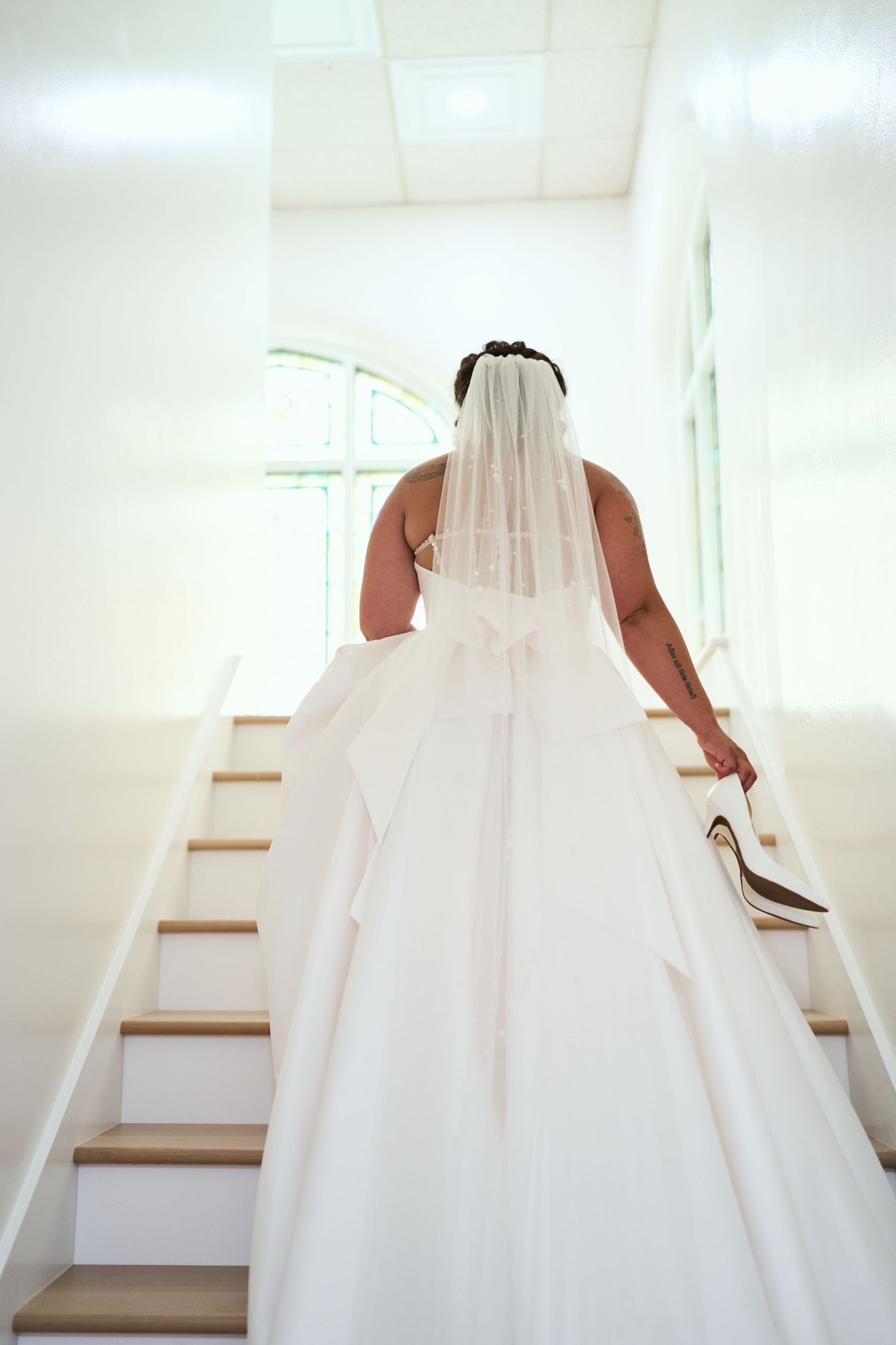 Back view of a bride in a classic white wedding gown ascending wooden stairs.