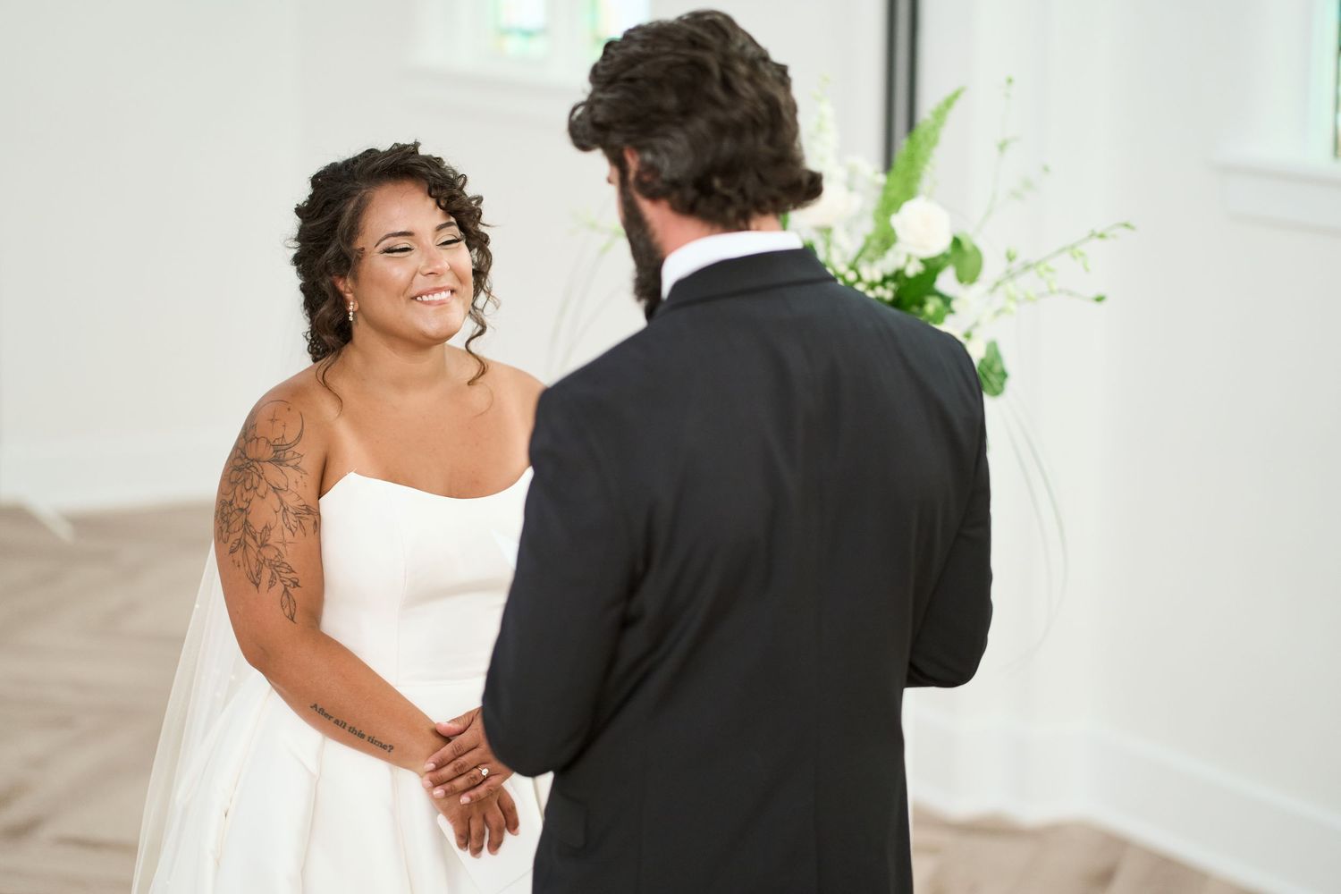 A sweet wedding moment between two people exchanging vows in a bright, modern venue with white floral arrangements.