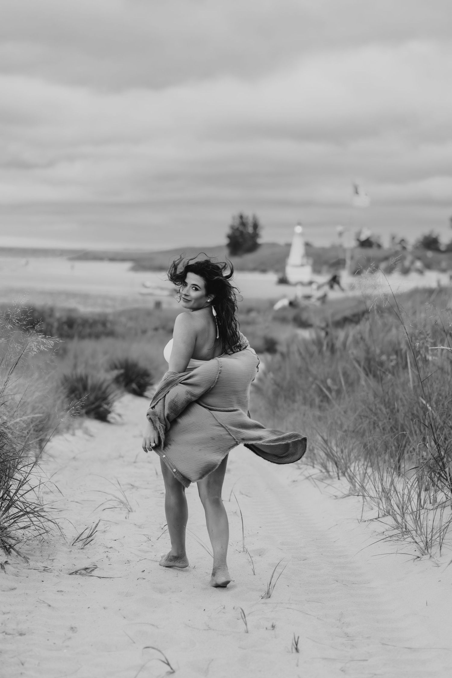 Woman in flowing dress poses near lighthouse on sandy coastal path in black and white photo.