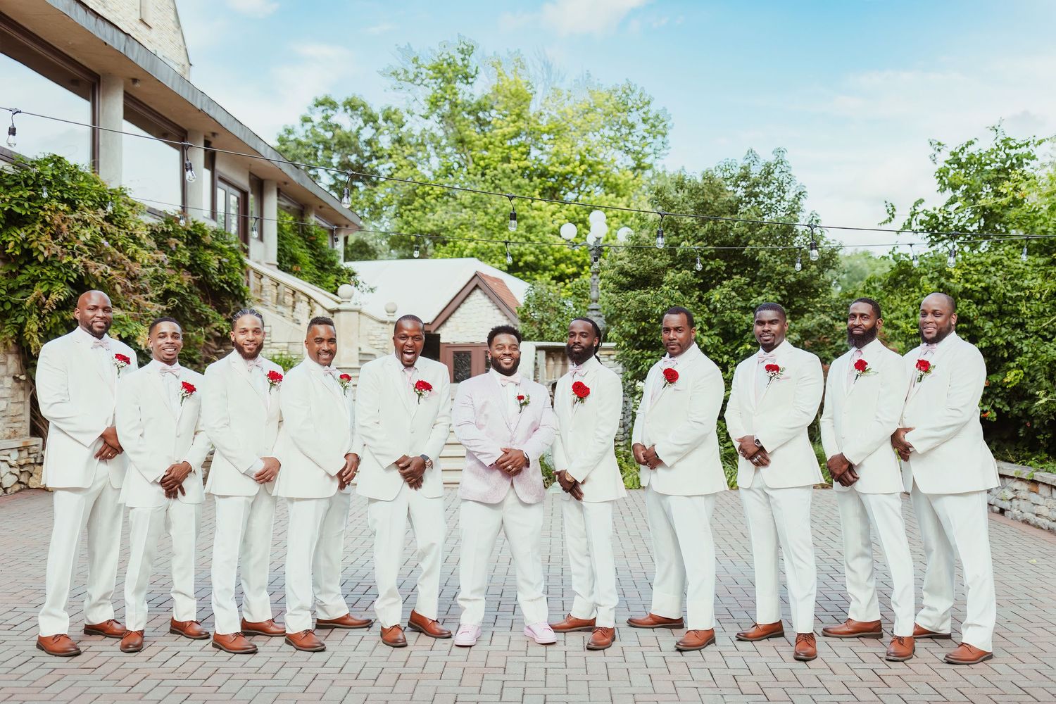 Groomsmen lineup wearing crisp white suits and brown shoes for outdoor wedding photo.
