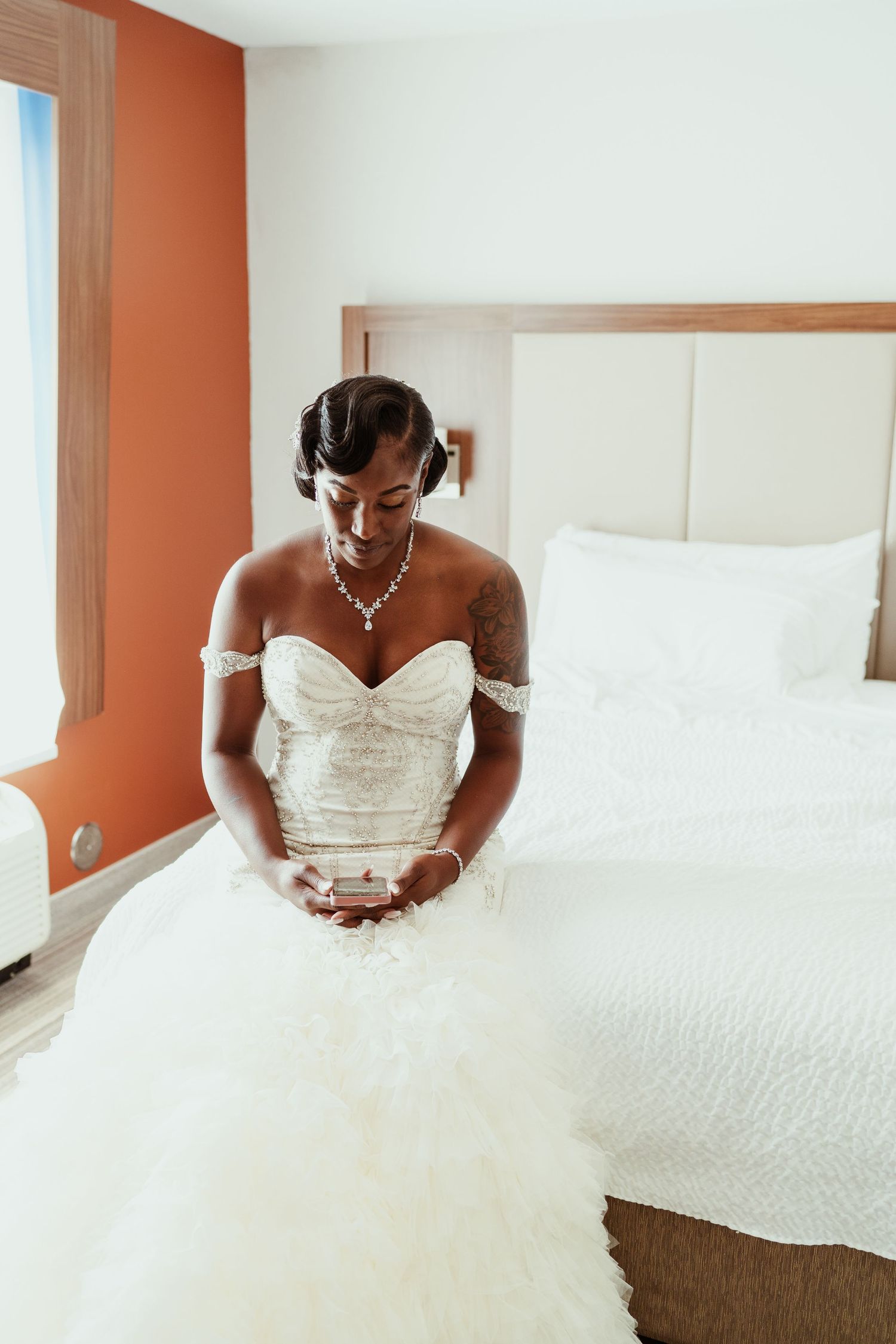Bride in a strapless white wedding gown with off-shoulder details sits peacefully on a hotel bed.