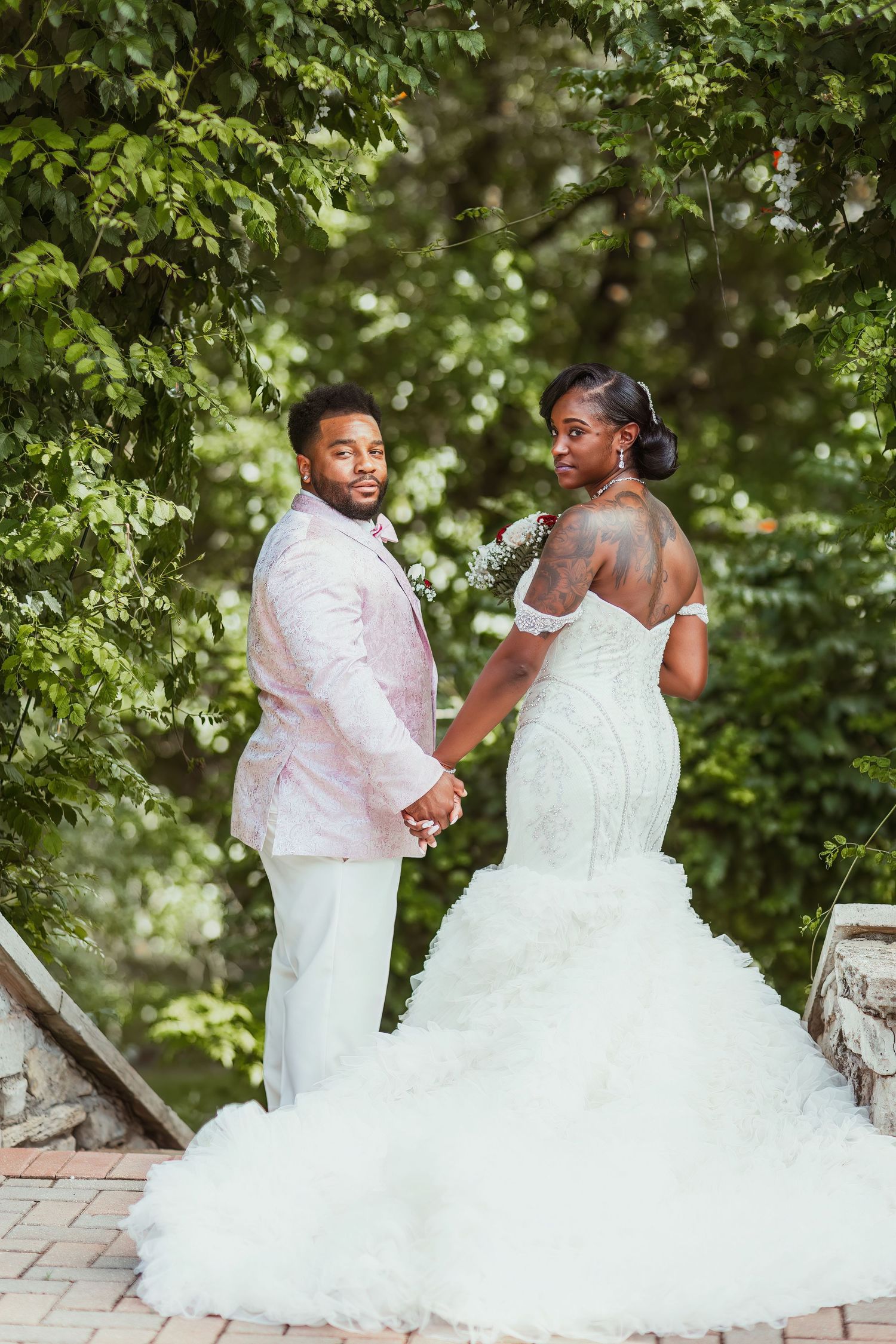 Couple in wedding attire poses on stone steps surrounded by lush greenery.