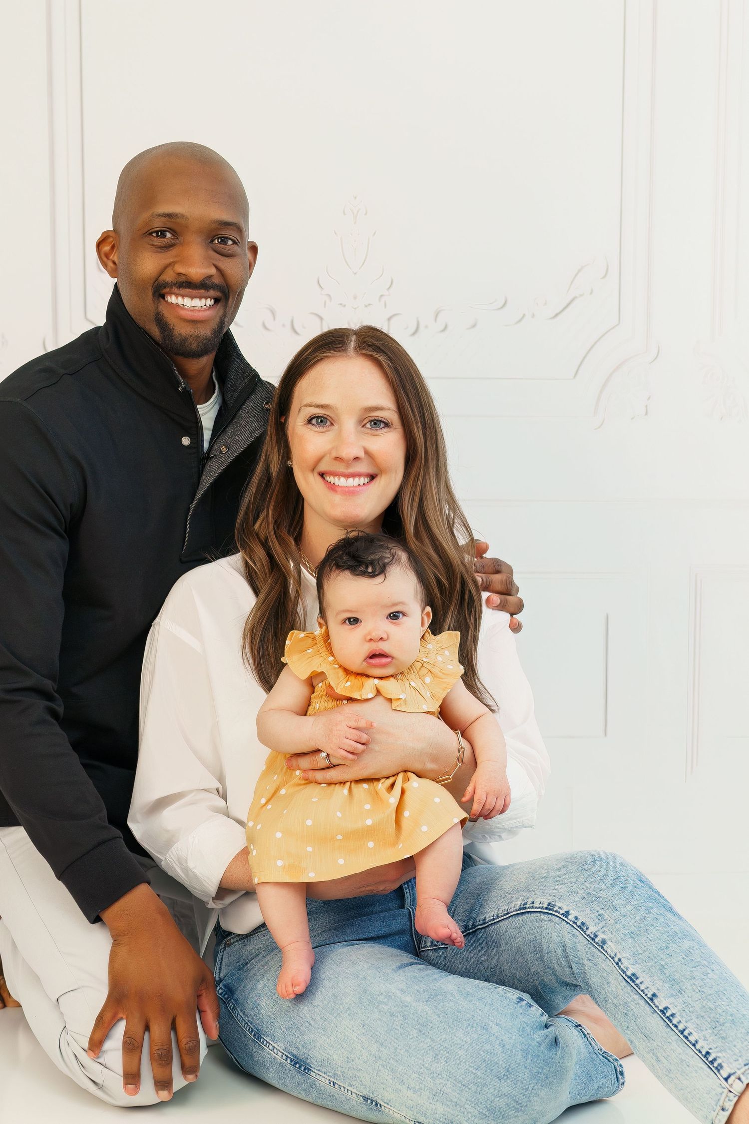 A family of three sits together on a white background in matching cream colored sweaters and jeans.