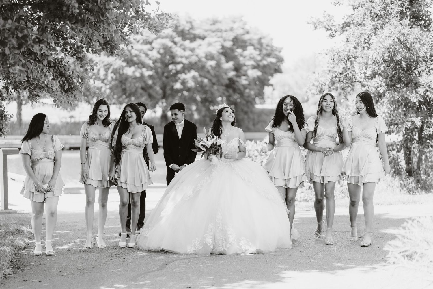 Black and white wedding photo showing a bride in a ballgown with bridesmaids in short dresses on a tree-lined path.