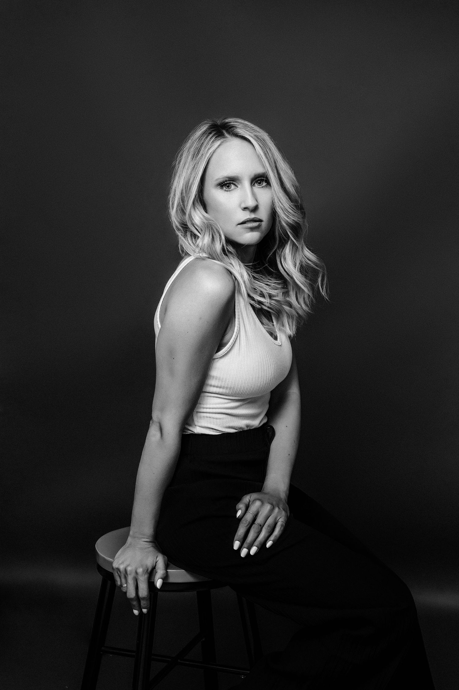 Black and white studio portrait of a woman in a white tank top and dark skirt sitting on a stool against dark background.