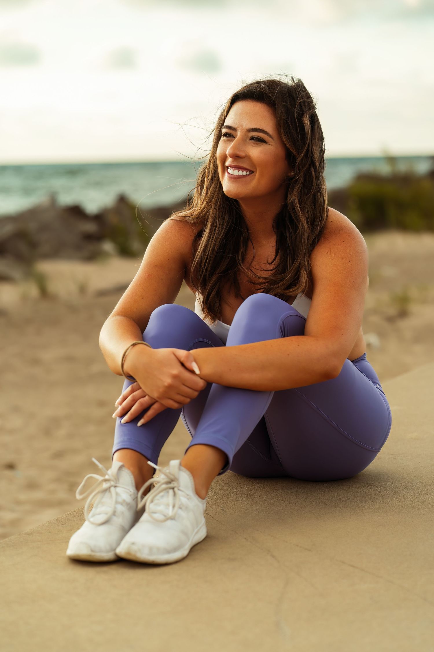 Woman in purple athletic wear smiles while sitting on beach during sunset.