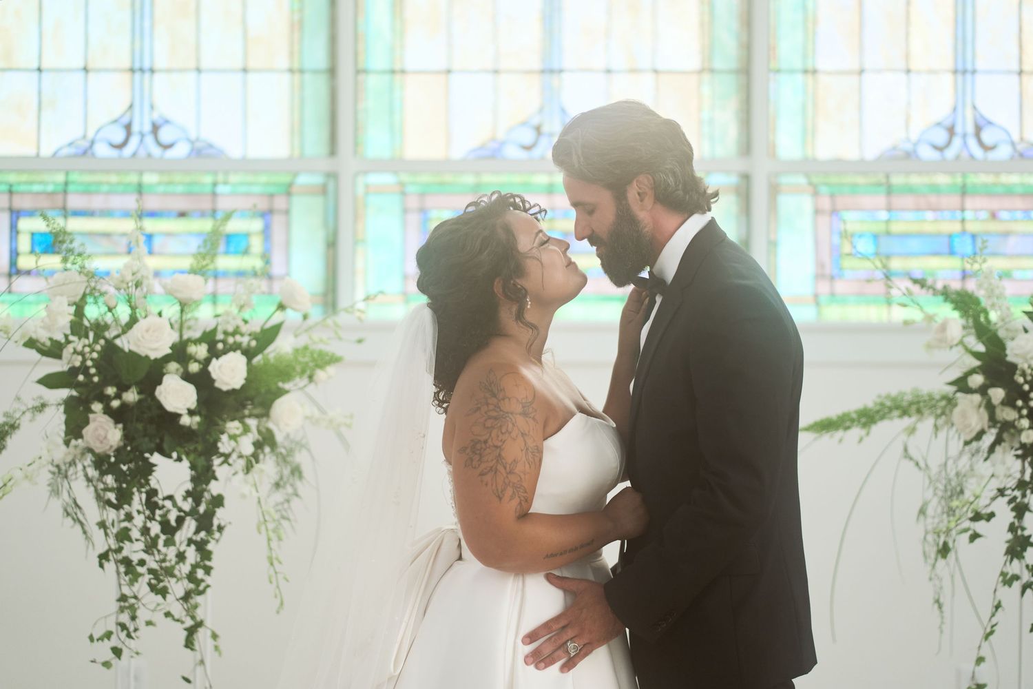 Romantic wedding portrait with sunlight streaming through stained glass windows and white floral arrangements.