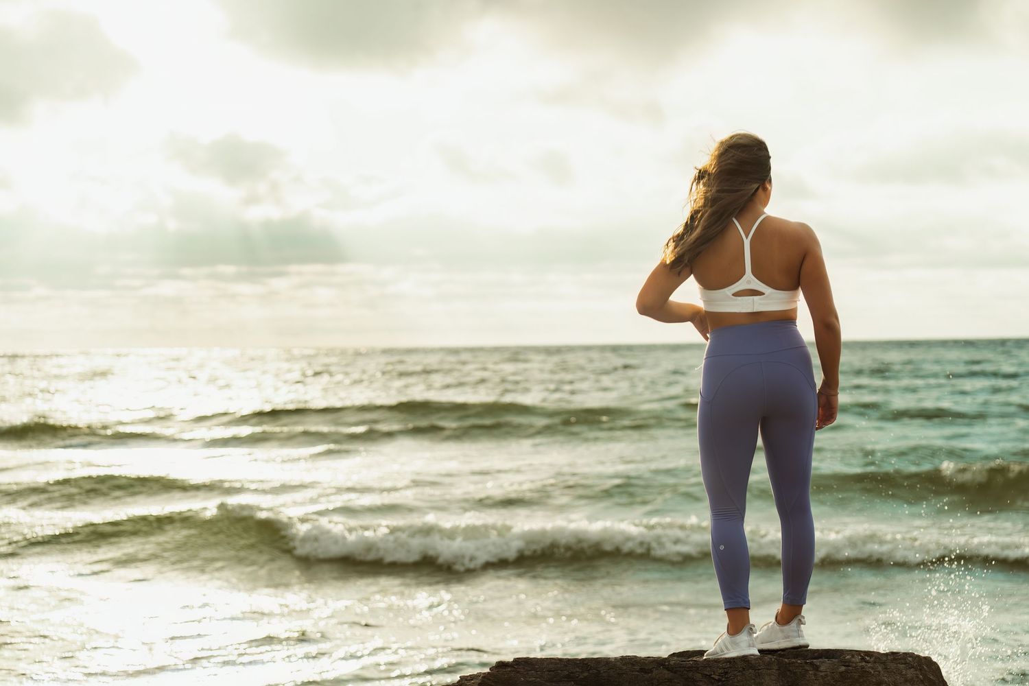 Silhouette of woman in workout clothes standing on rocks by ocean at sunset.
