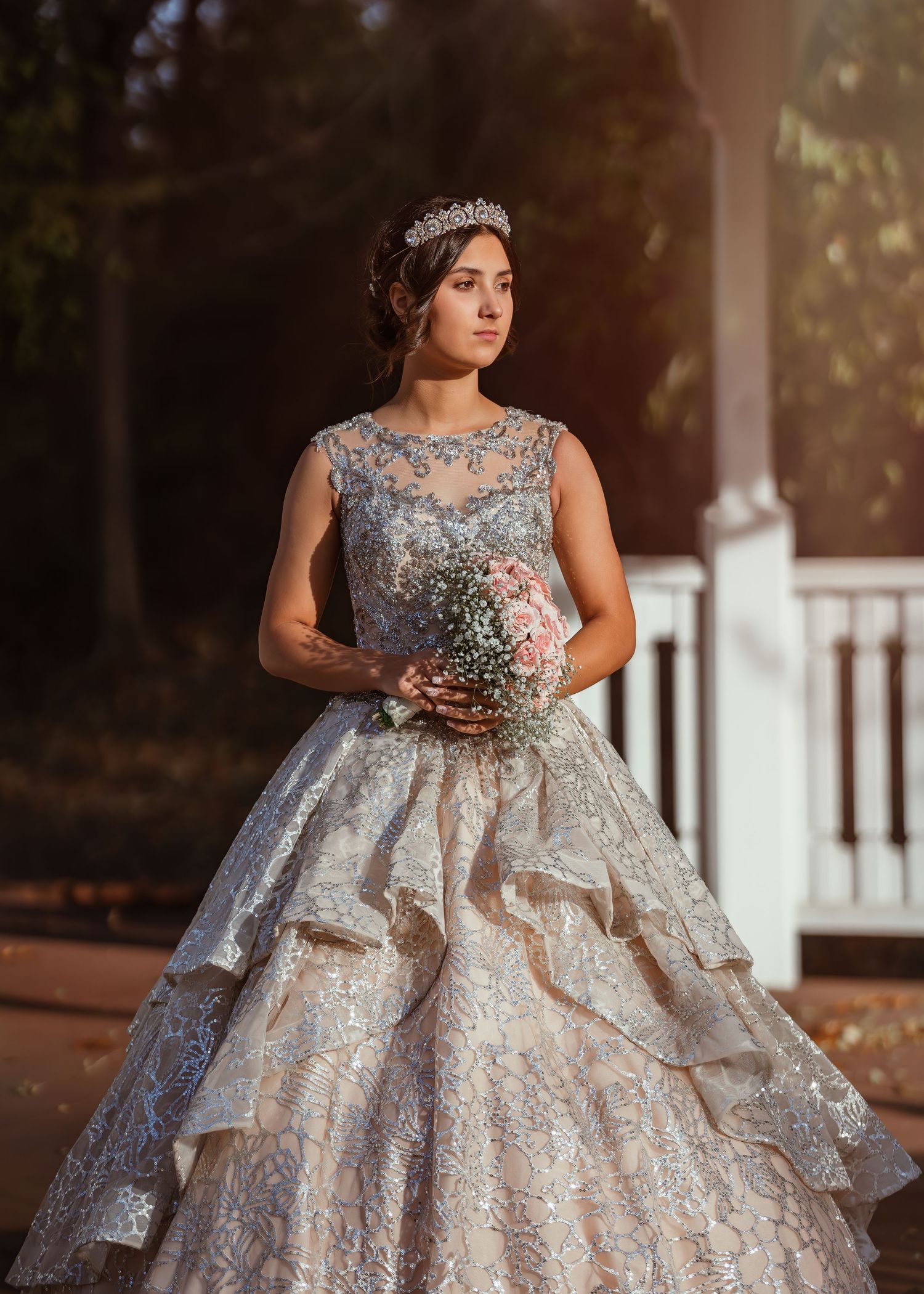 Elegant bride in a sparkly ball gown with tiered skirt holding a bouquet against a sunlit gazebo backdrop.