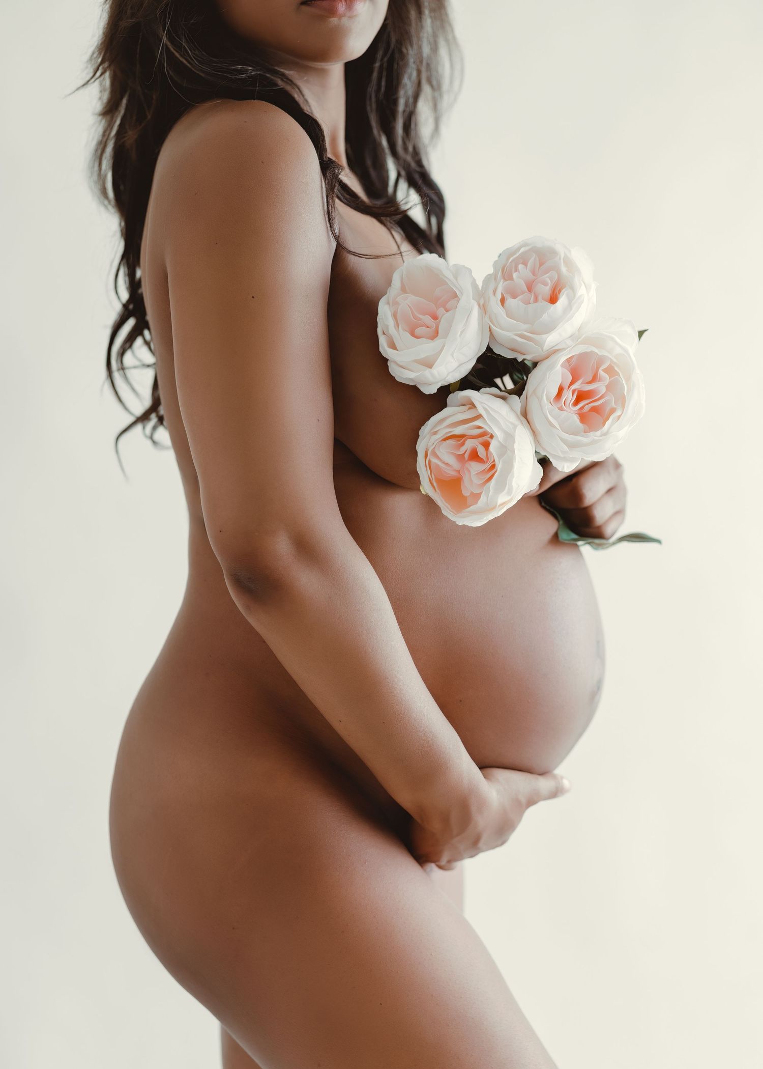 Artistic maternity photo with nude silhouette holding pale pink roses against white background.