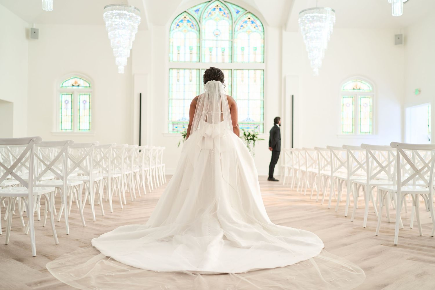 Sunlit wedding chapel interior with white cross-back chairs and dramatic train of wedding dress.