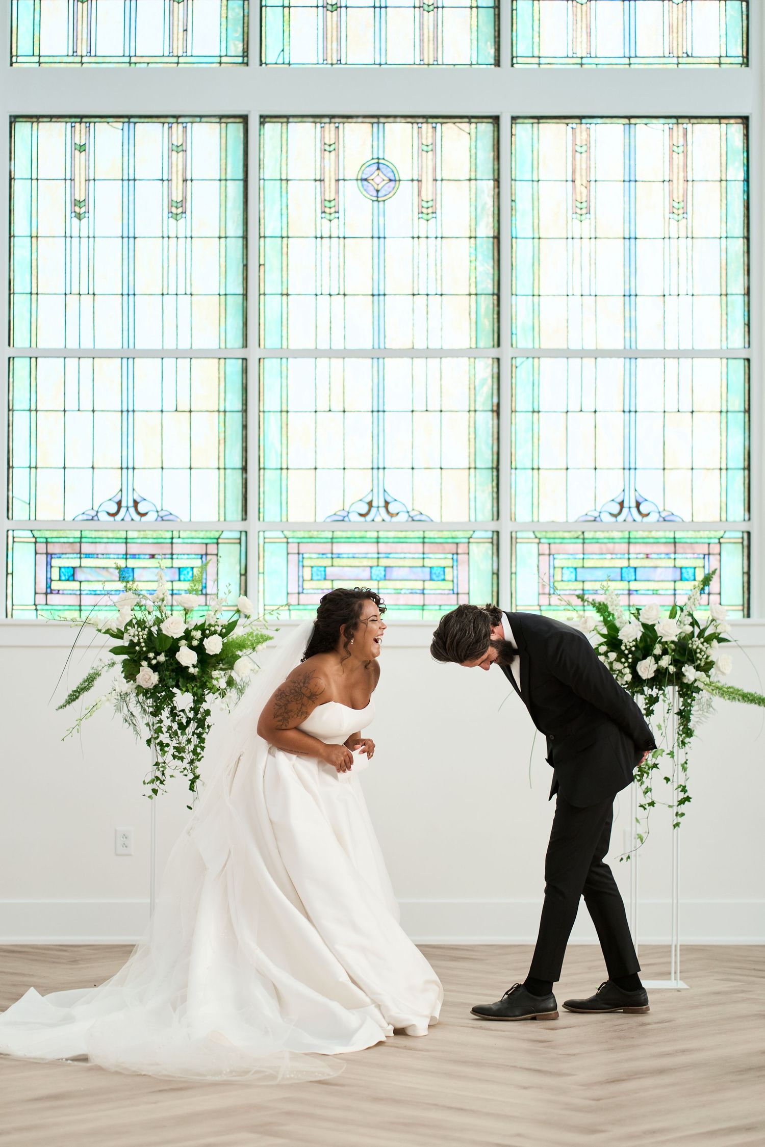 A lighthearted moment between newlyweds against stunning stained glass windows and white floral decorations.