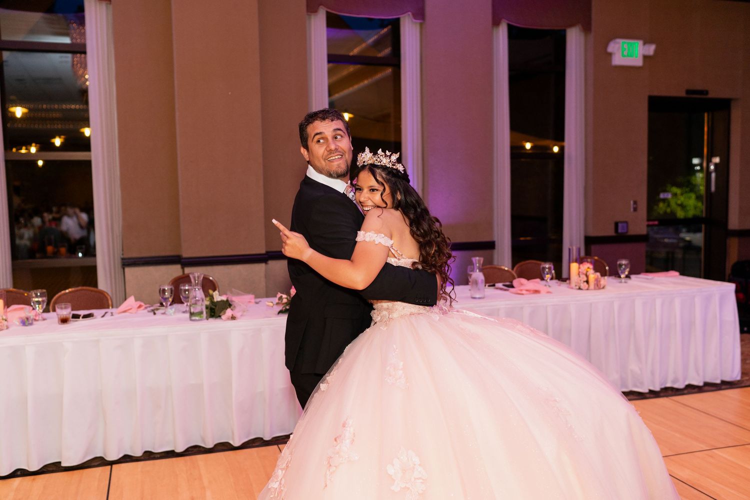 A couple shares their first dance at their wedding reception in a decorated ballroom with white-draped tables.