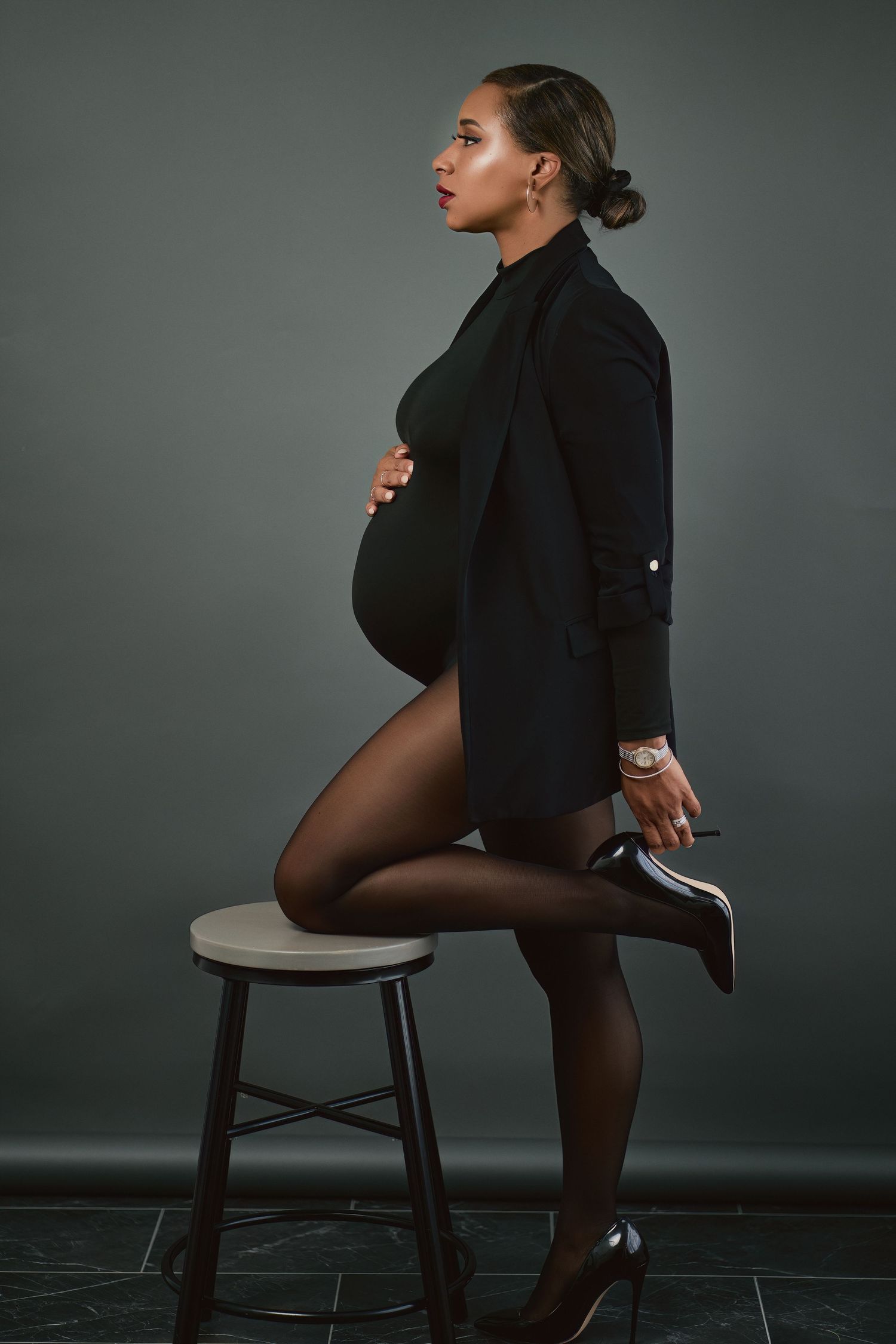 A figure in black attire poses on a chair against a dark gray backdrop in an artistic studio portrait series.
