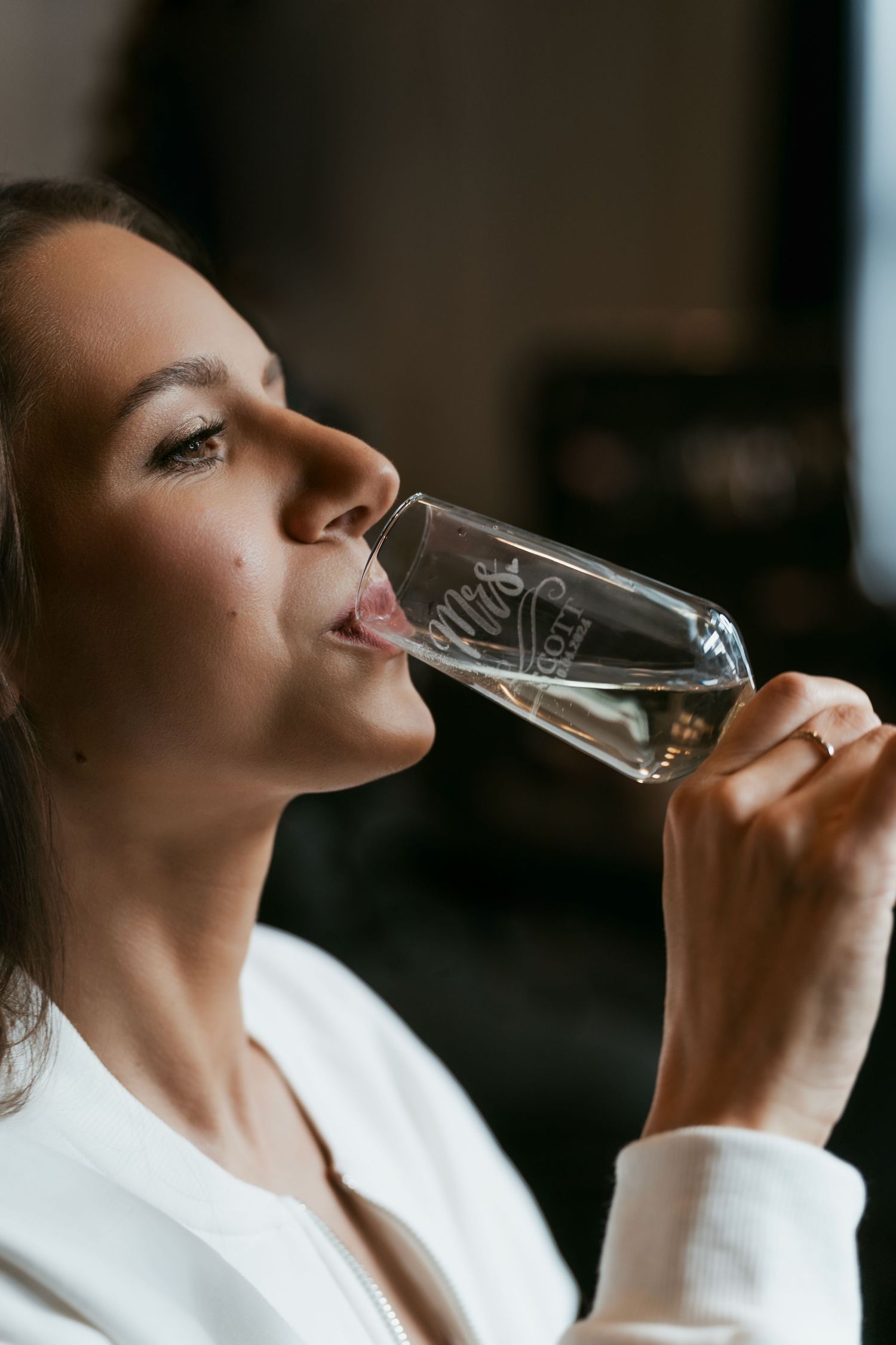 Side profile view of someone in a white robe sipping from an elegant wine glass in a dimly lit setting.