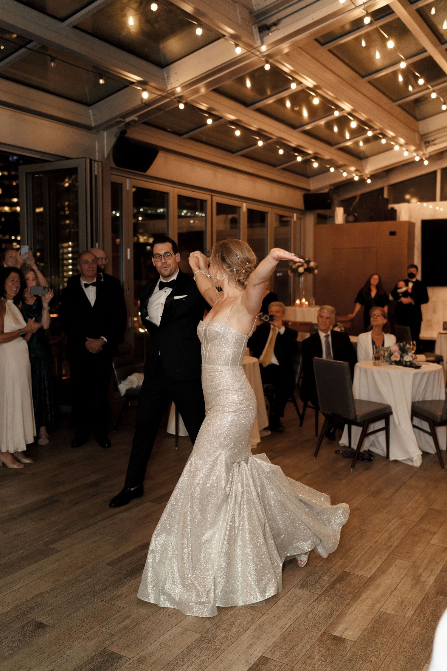 Wedding guests dance and celebrate on a wooden dance floor under warm lighting at an indoor reception venue.