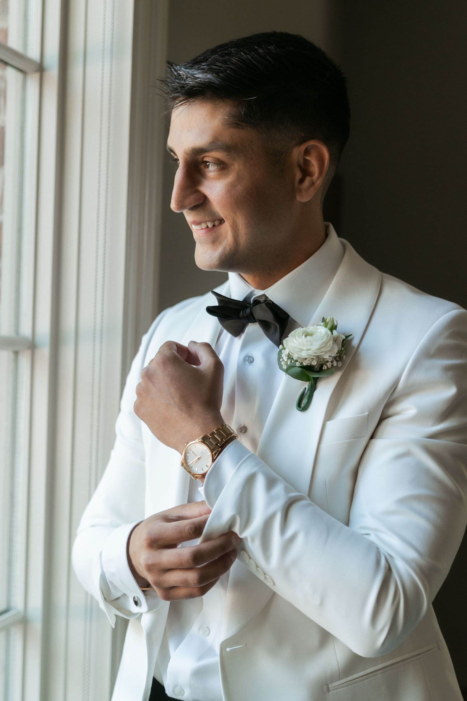 Groom in white tuxedo adjusts his watch and boutonniere while getting ready near a window.