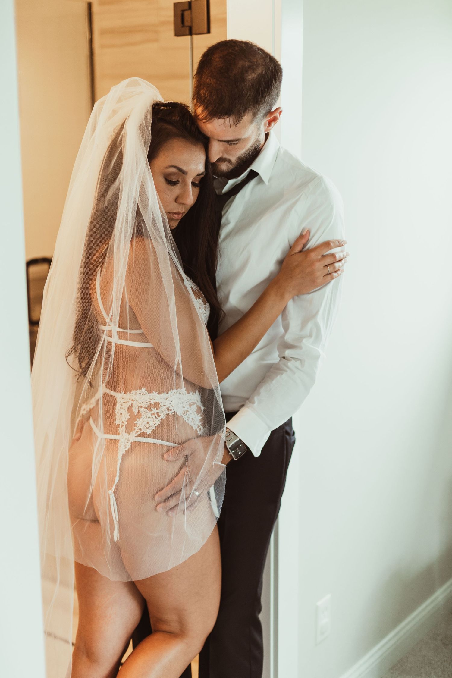 Intimate wedding boudoir photo of couple embracing in white formal wear and lingerie.