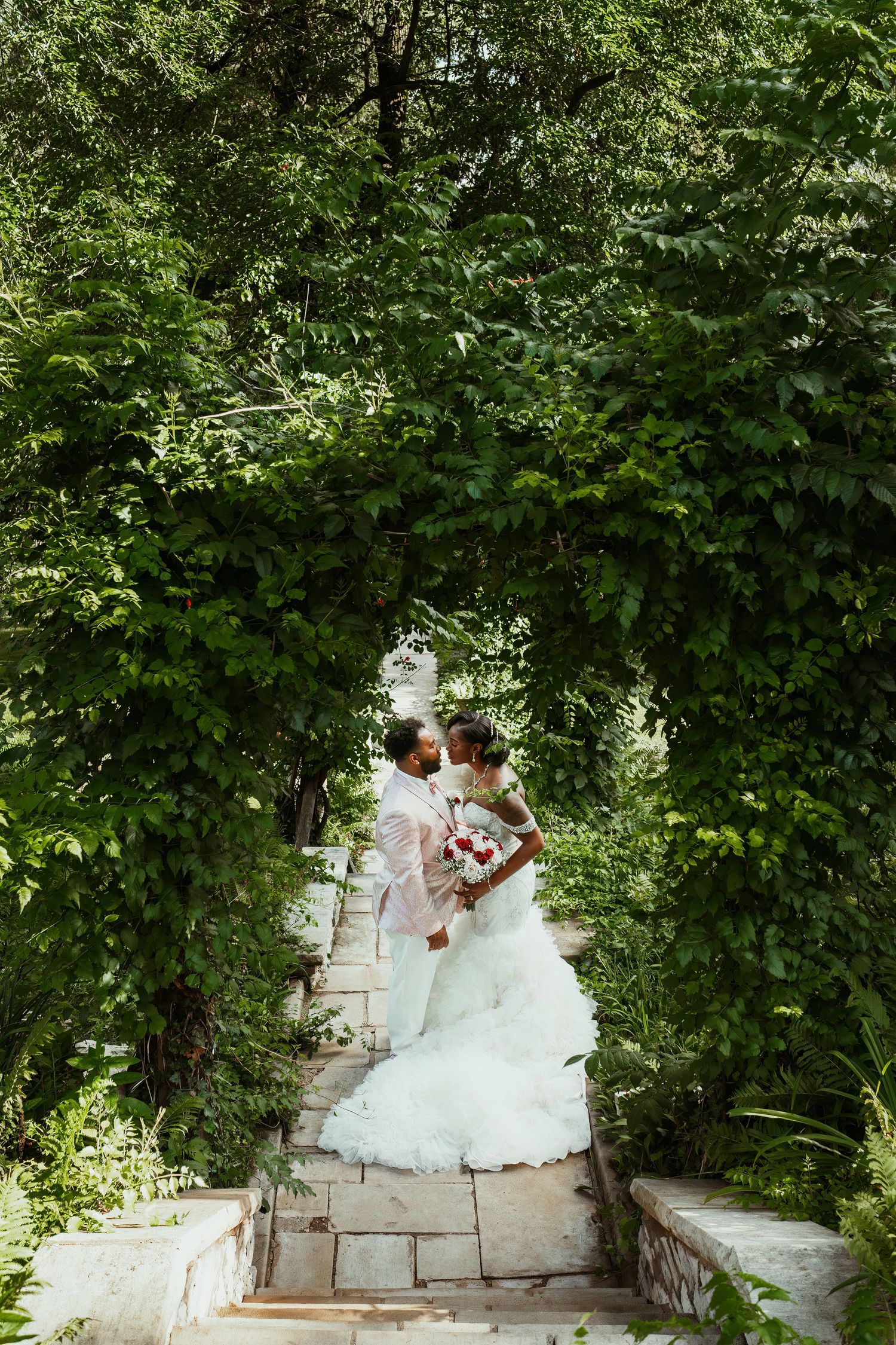 A couple descends stone stairs through a lush green garden archway in wedding attire.