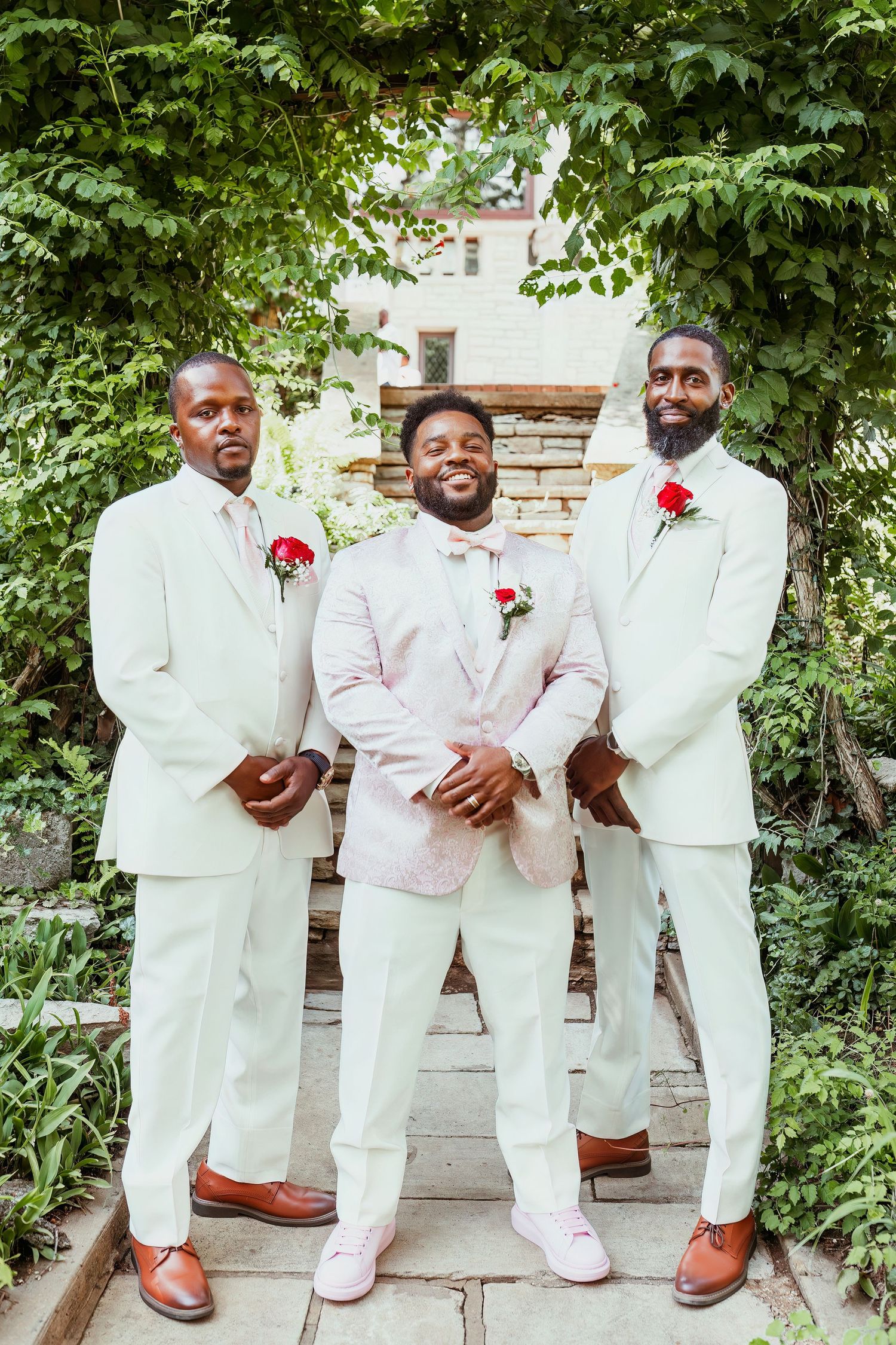 Three groomsmen in elegant white and light pink suits with red boutonnieres pose on outdoor stone steps.