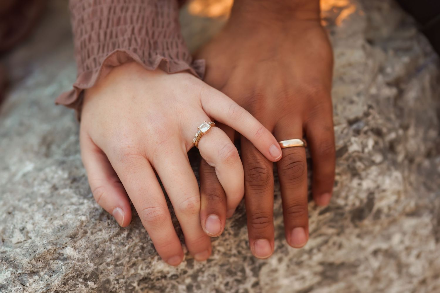 Intimate detail shot of intertwined hands showing wedding rings on stone surface.