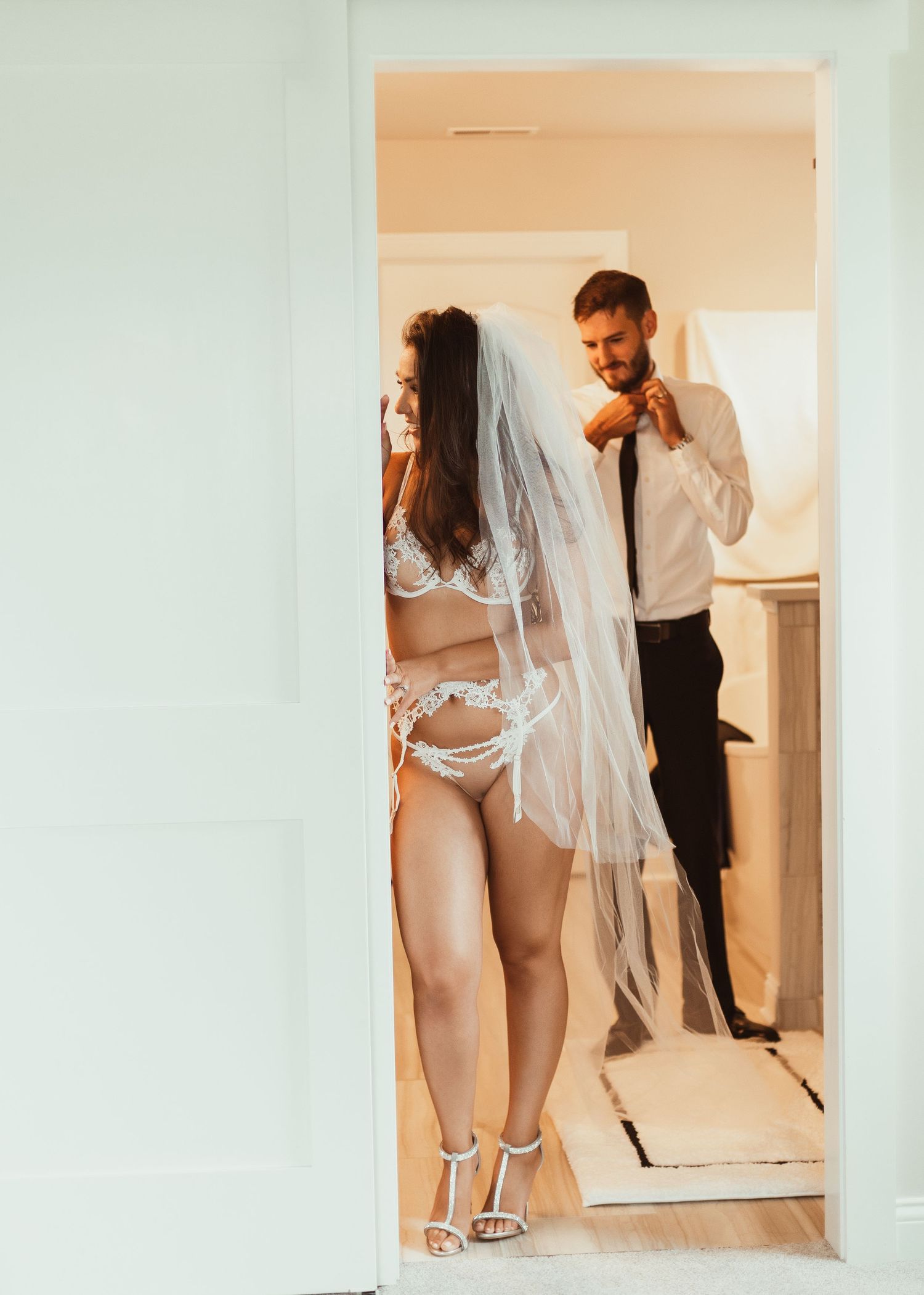 A couple poses in a mirror wearing wedding attire, with the bride in a white veil and lingerie and groom in a suit.