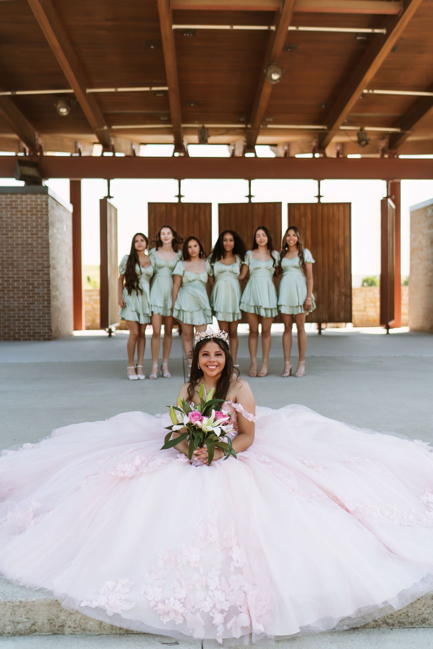 A bride in a pink ball gown poses with bridesmaids wearing mint dresses under a wooden pavilion.