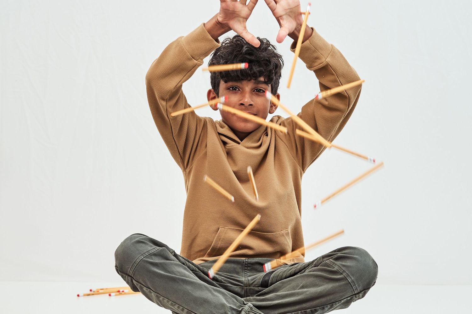 A person in a tan hoodie and gray pants sits cross-legged while gesturing playfully with their arms above their head.