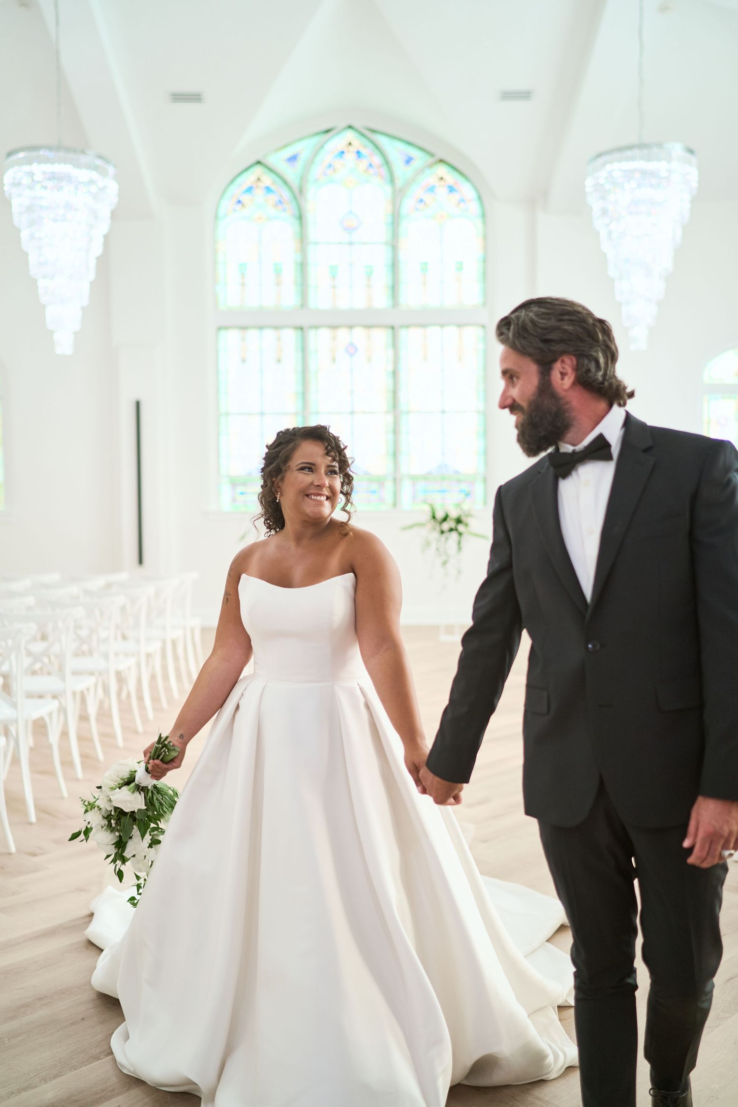A couple in wedding attire walk hand in hand inside a bright chapel with stained glass windows and white chairs.