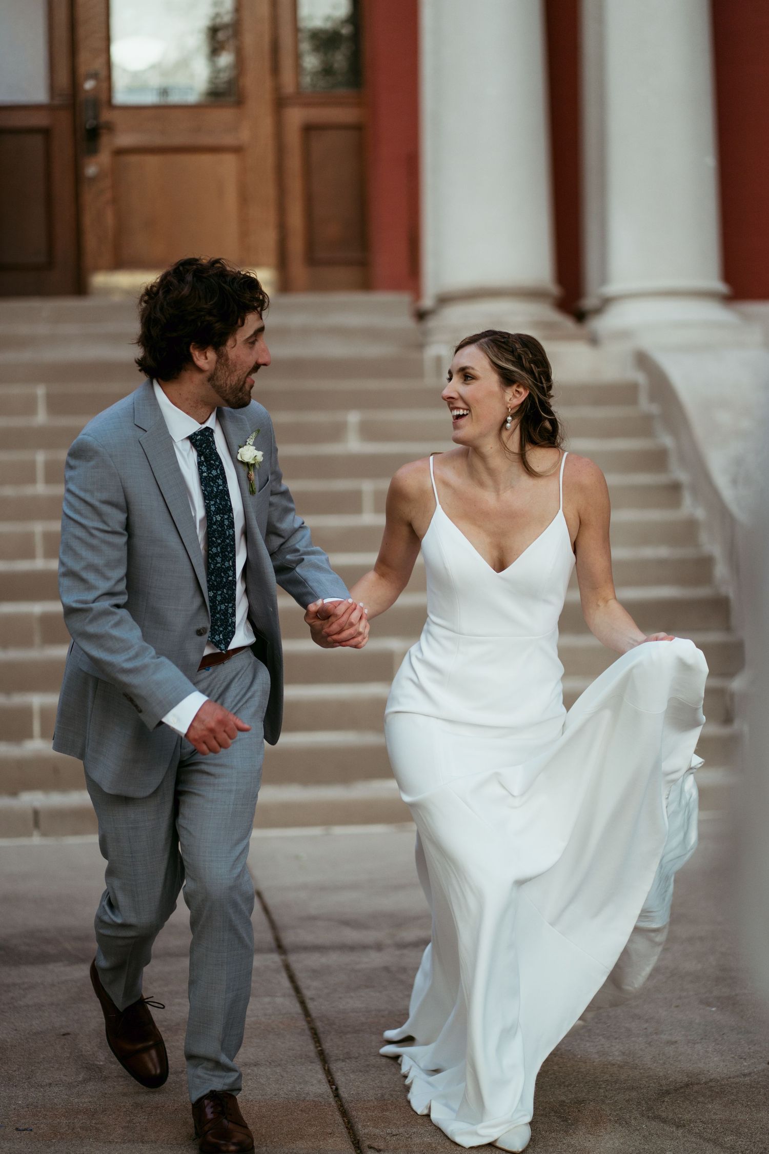 A couple in wedding attire playfully runs together on stone steps, the bride's white dress flowing in motion.