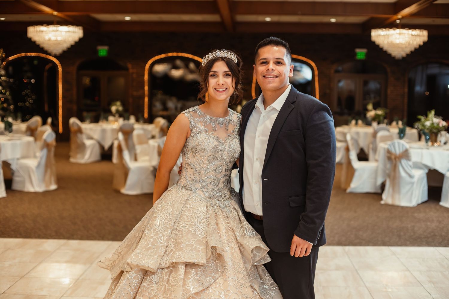 A couple stands together in an elegant ballroom with crystal chandeliers and white-draped chairs in the background.
