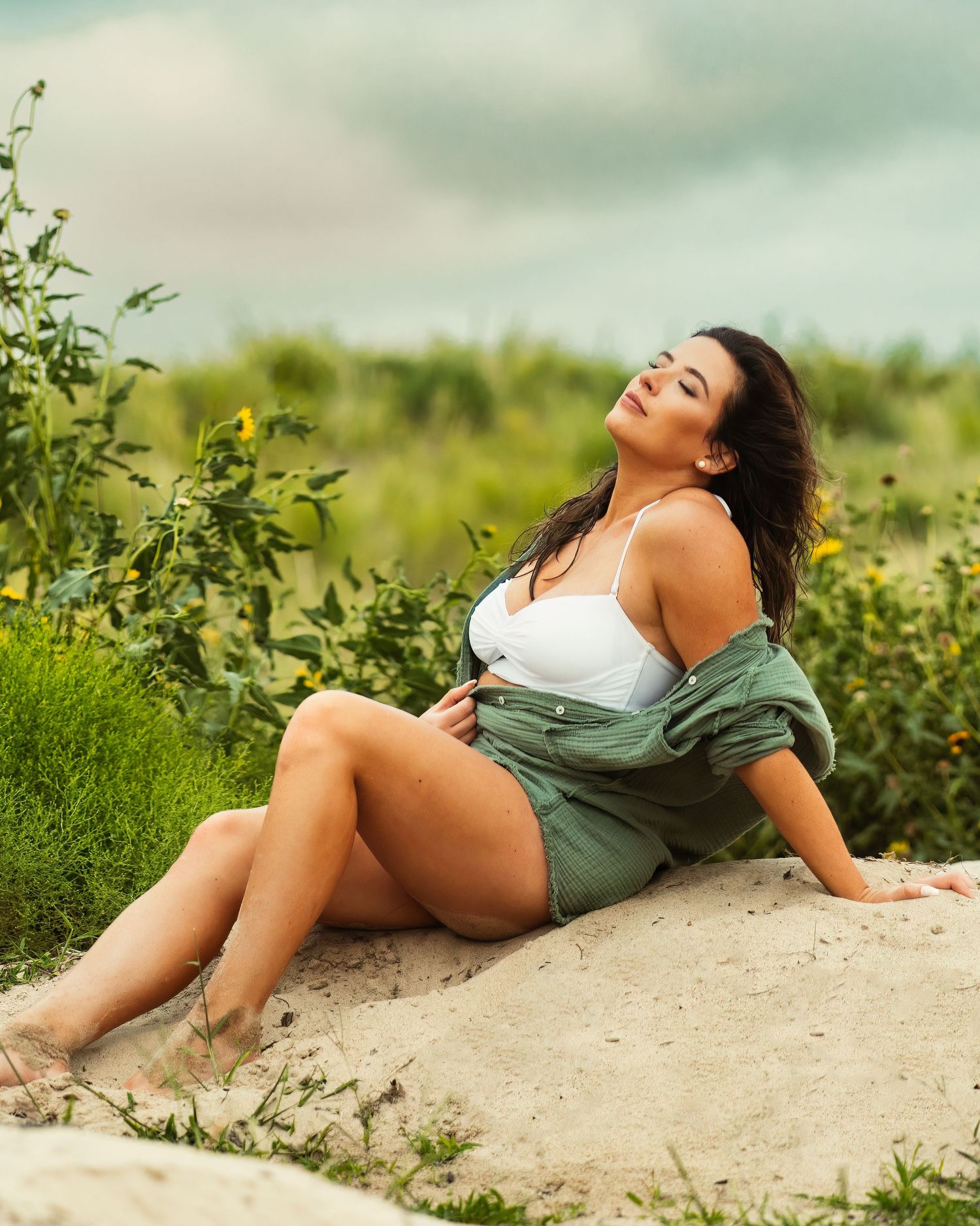 Woman in swimwear and green jacket reclines on sandy ground surrounded by summer vegetation.
