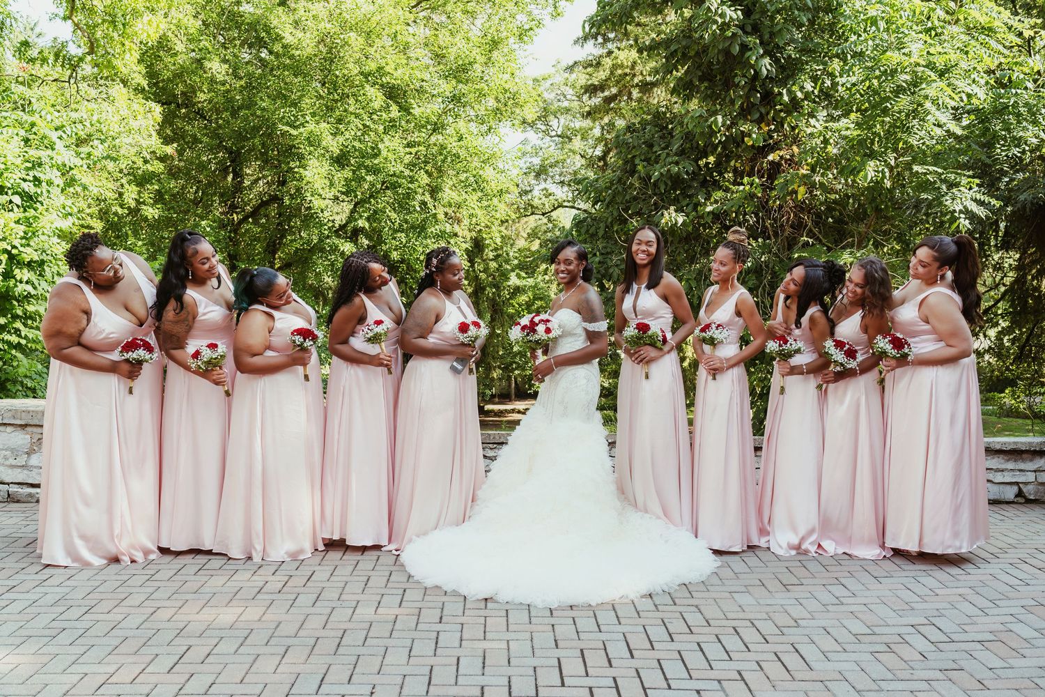 Wedding party photo with bridesmaids in pink gowns flanking bride's white dress outdoors.