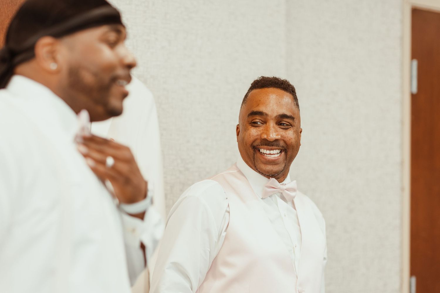 Two people in white formal attire sharing a joyful moment while getting ready.