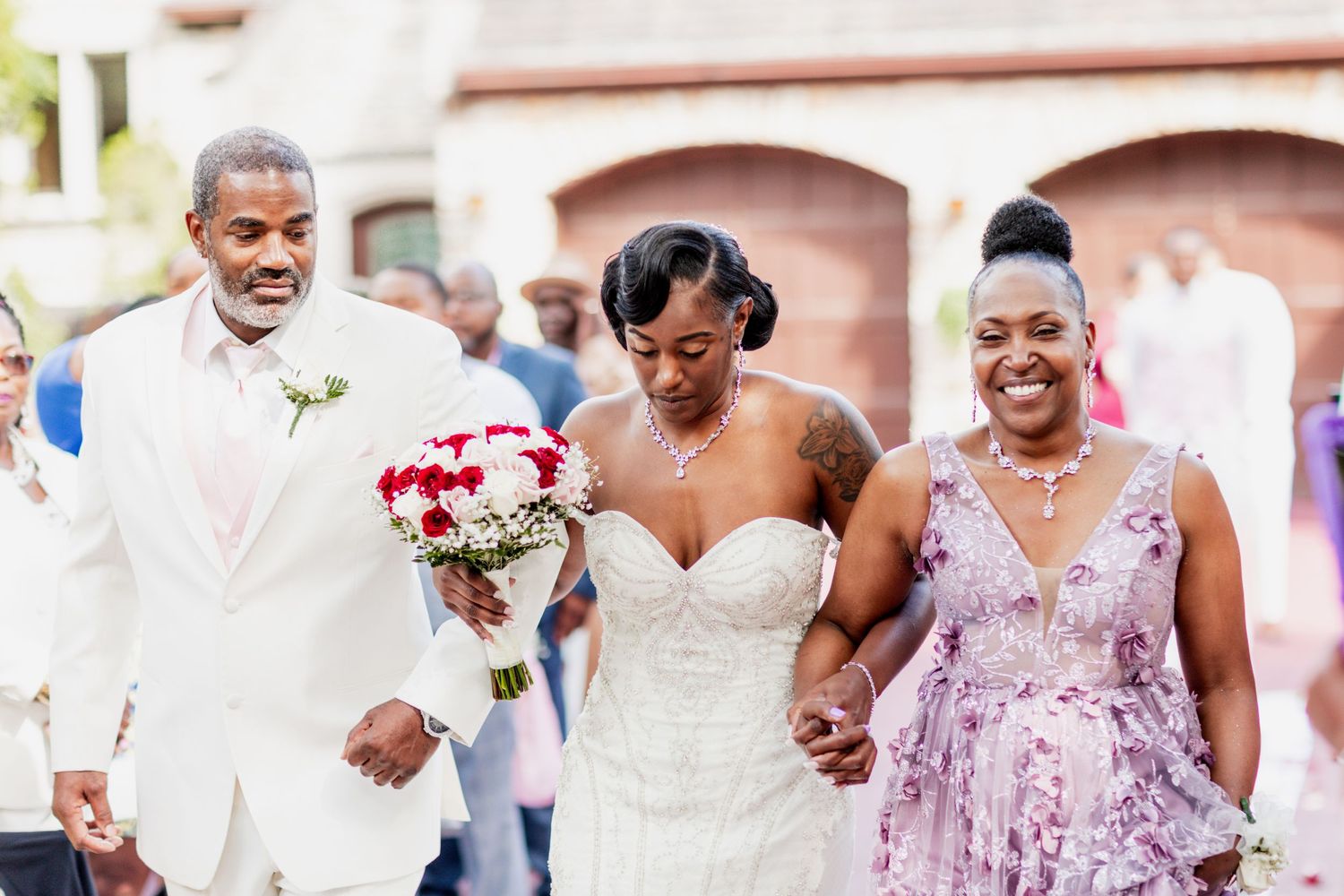 Three wedding attendees walk together outdoors, with the middle person holding a red and white bouquet.