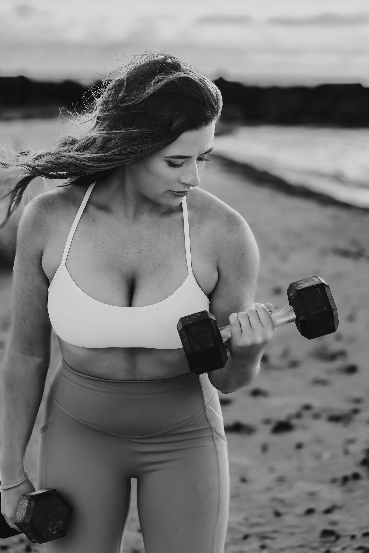 Woman in activewear exercises with dumbbells during beach workout in monochrome.
