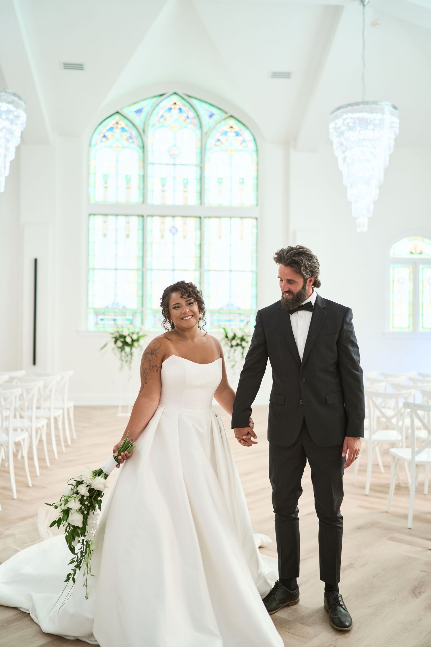 A newlywed couple walks together at a sunlit chapel venue decorated with white chairs and elegant windows.