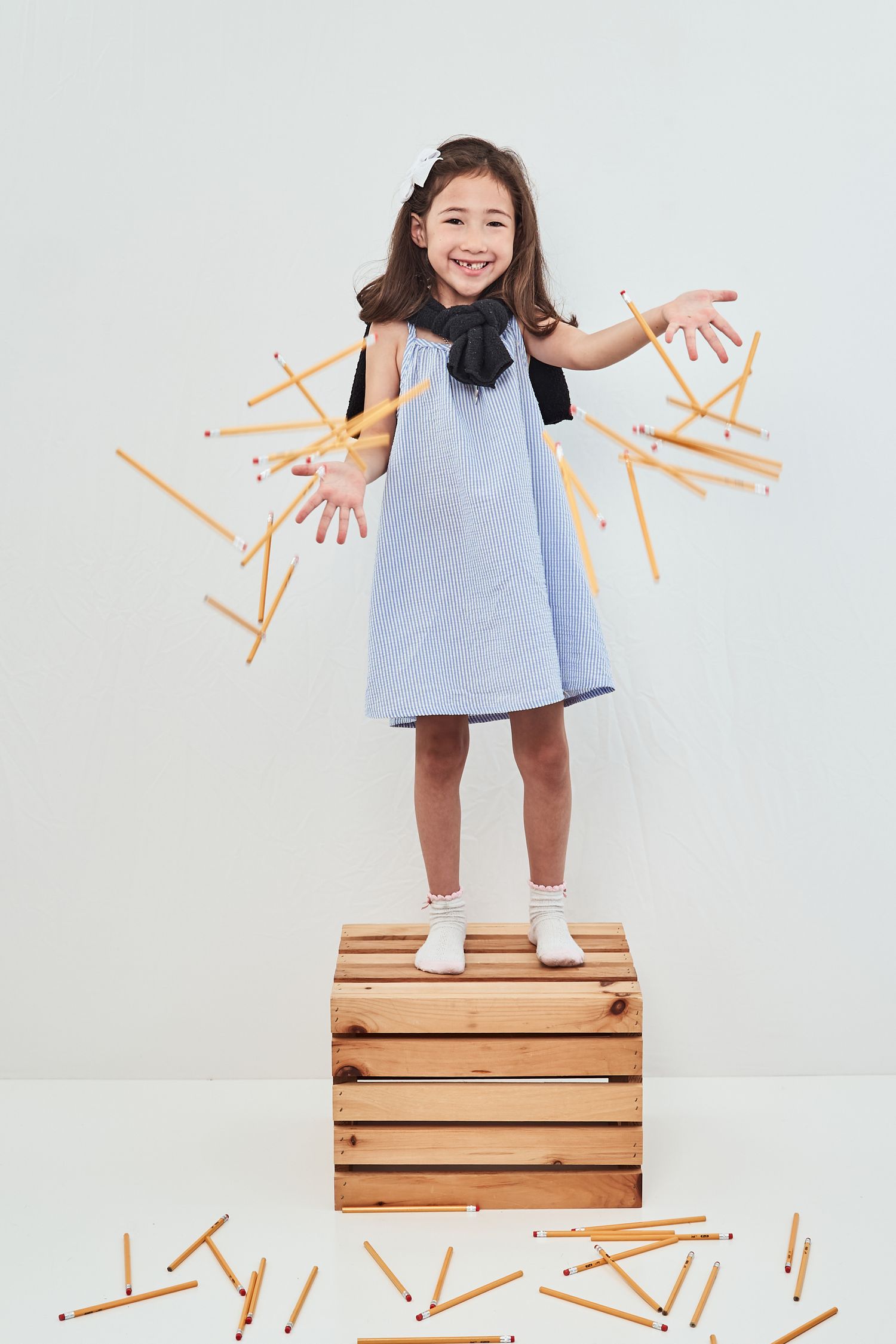 Young child in blue dress stands on wooden crate spreading arms while colorful pencils fall around them playfully.
