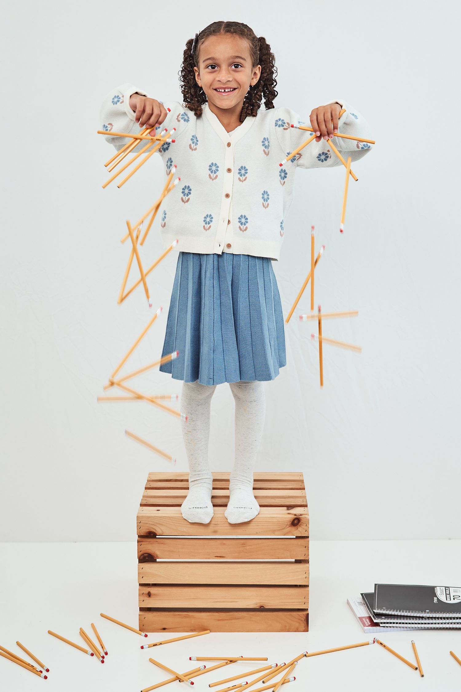Young child stands on wooden storage crate wearing blue dress with white pattern and tights against white background.