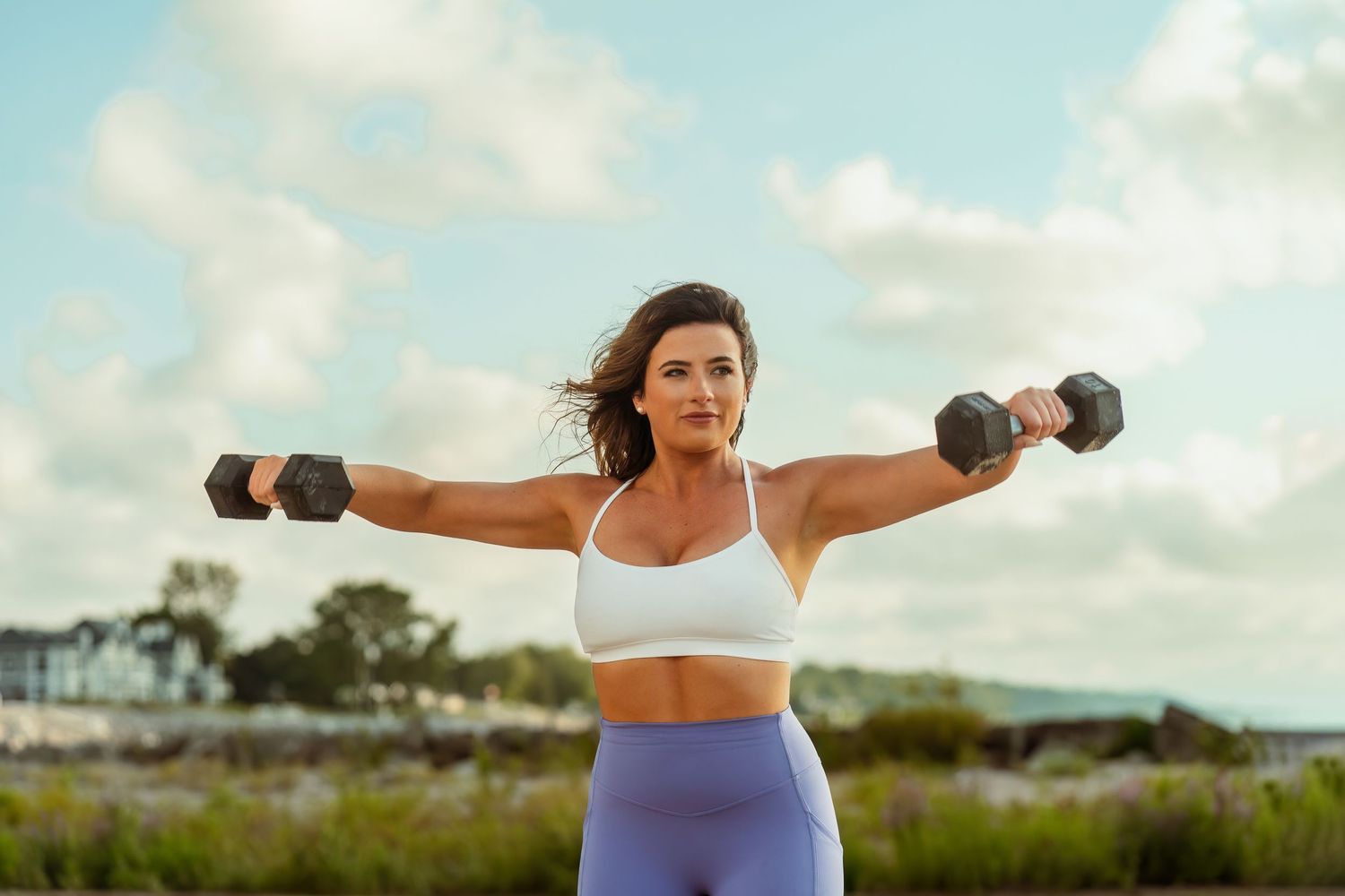 Fitness enthusiast performs lateral raises with dumbbells during outdoor beach workout in athletic wear.