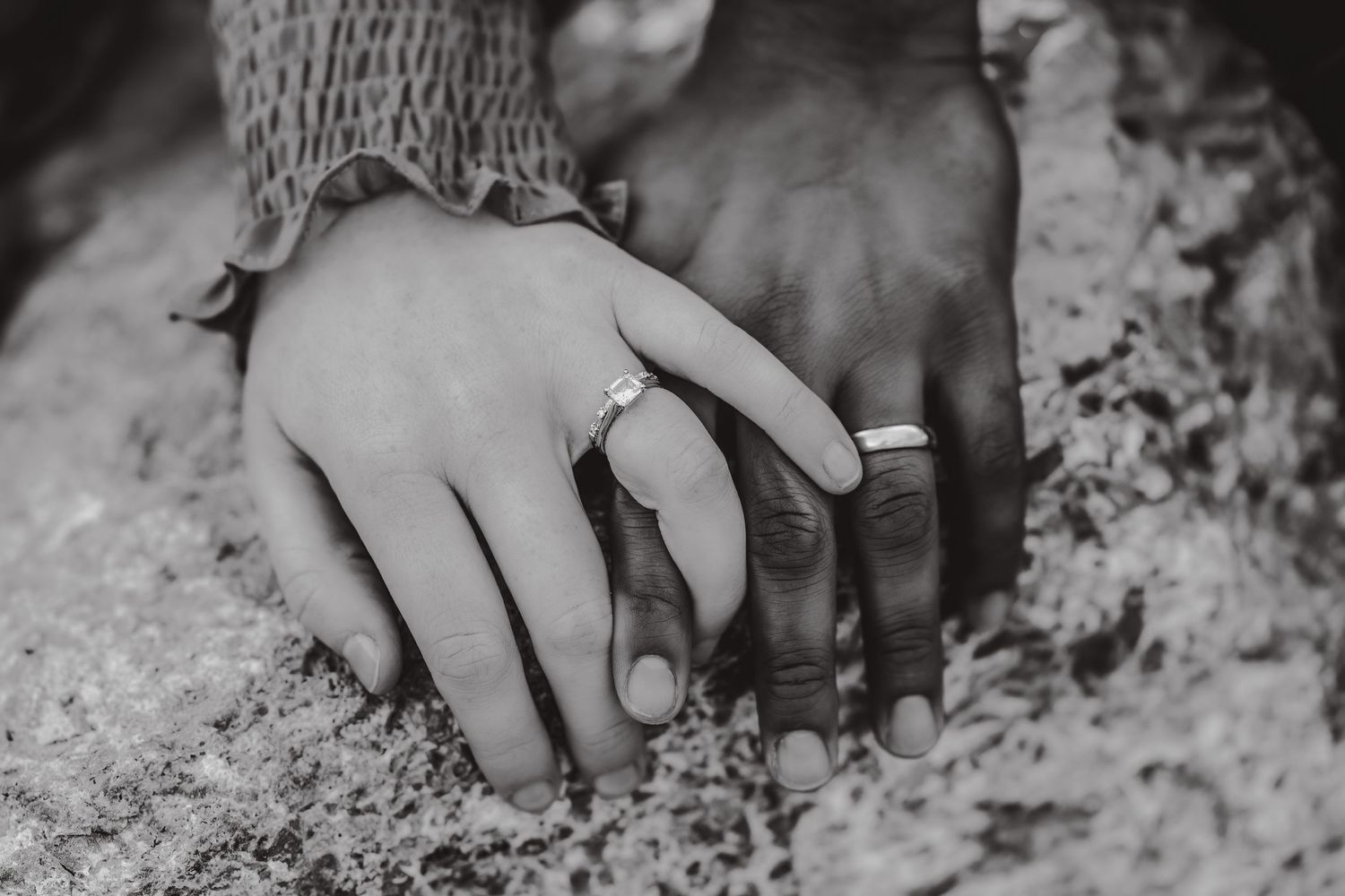 Close-up black and white shot of hands wearing wedding rings against textured stone.