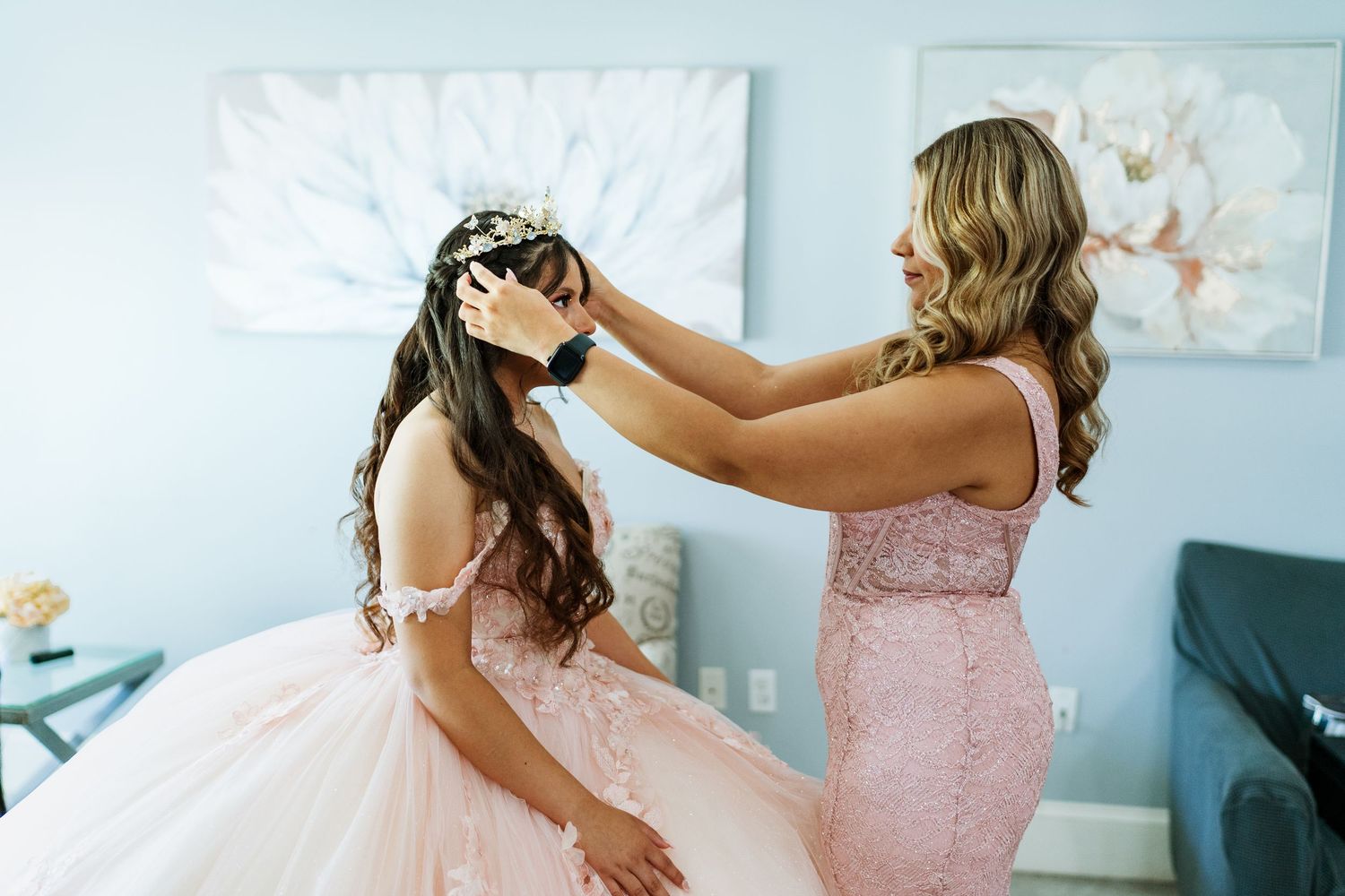 A bridesmaid in a pink dress helps adjust a tiara on a bride wearing a puffy white tulle wedding gown.